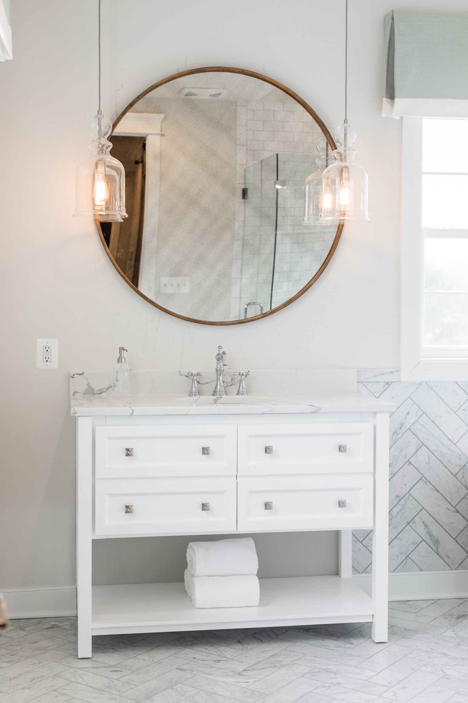 White bathroom vanity with round mirror, pendant lights, and stacked towels.