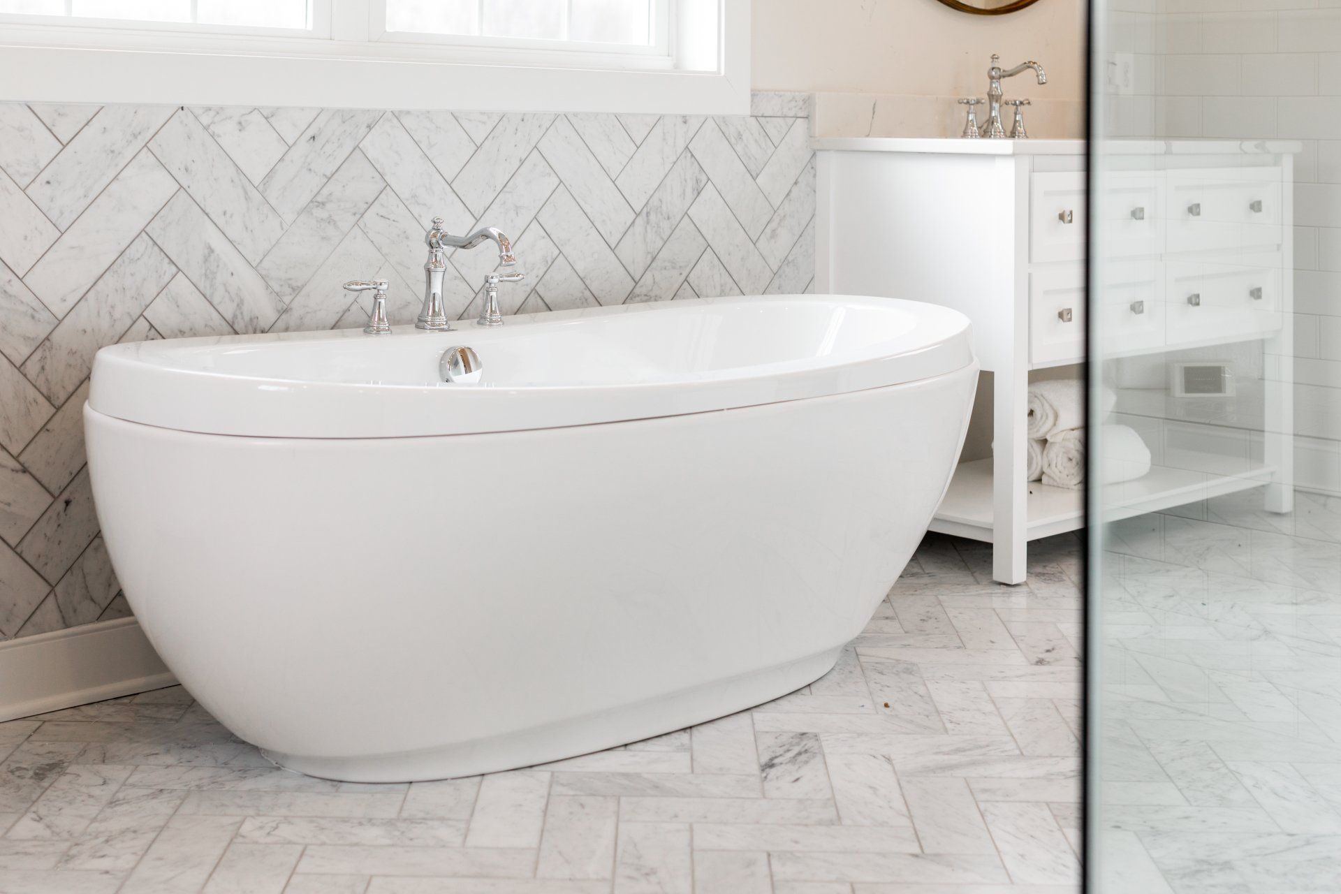 White oval bathtub in a bathroom with marble herringbone tile, a white vanity, and glass shower door.