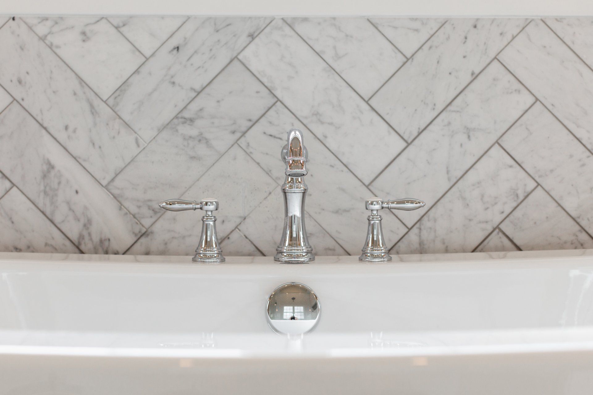 White bathtub with chrome faucet and herringbone marble backsplash.