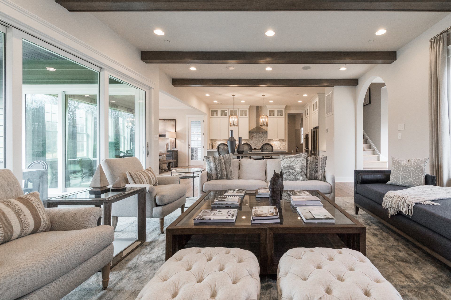 Spacious living room with neutral colors, large windows, open to kitchen area. Furnished with sofas, chairs, and ottoman.