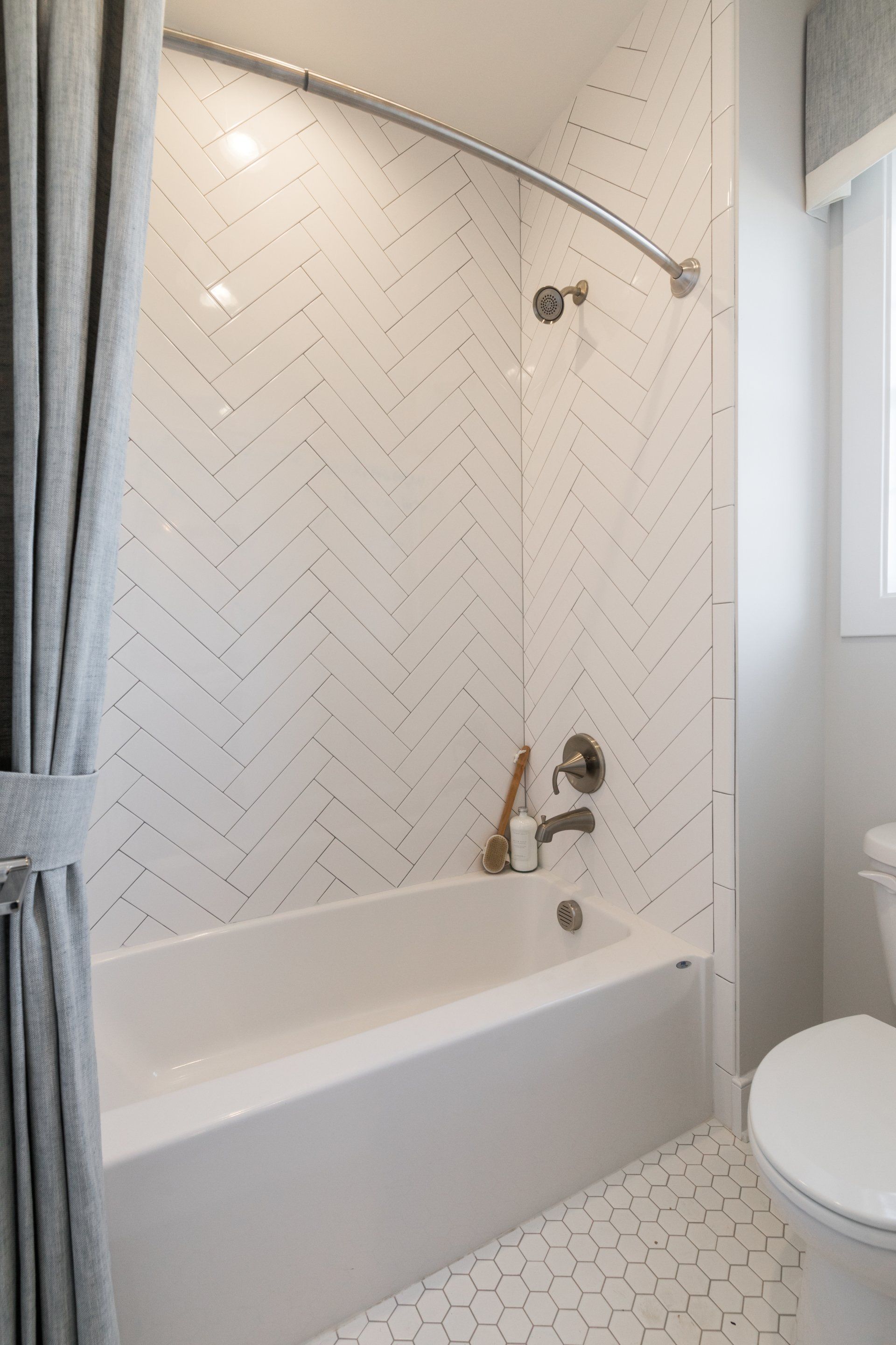 Bathroom with white tub, herringbone tile, silver showerhead, and gray shower curtain.