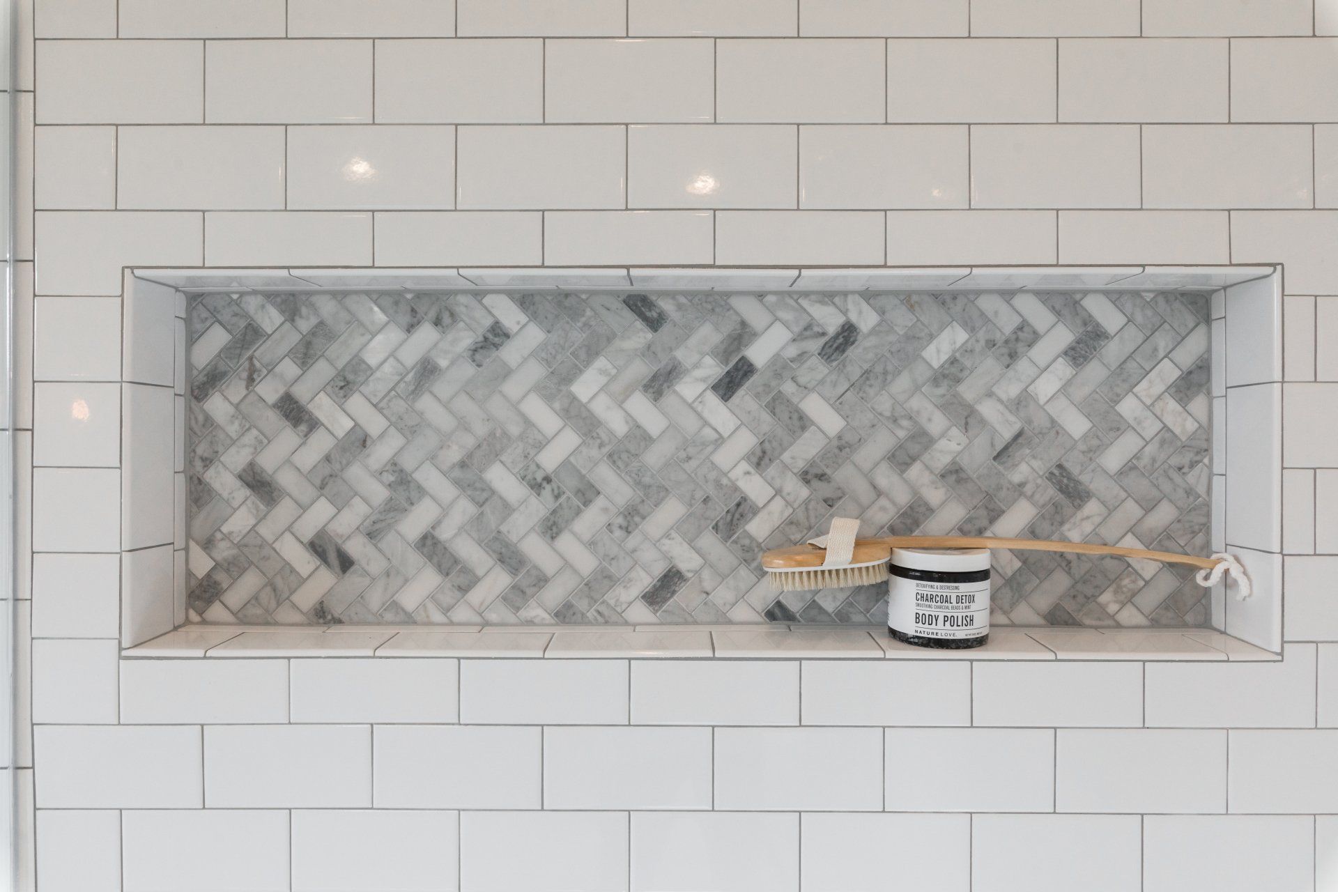 Recessed shower shelf with gray herringbone tile, white subway tile surround, and a jar.