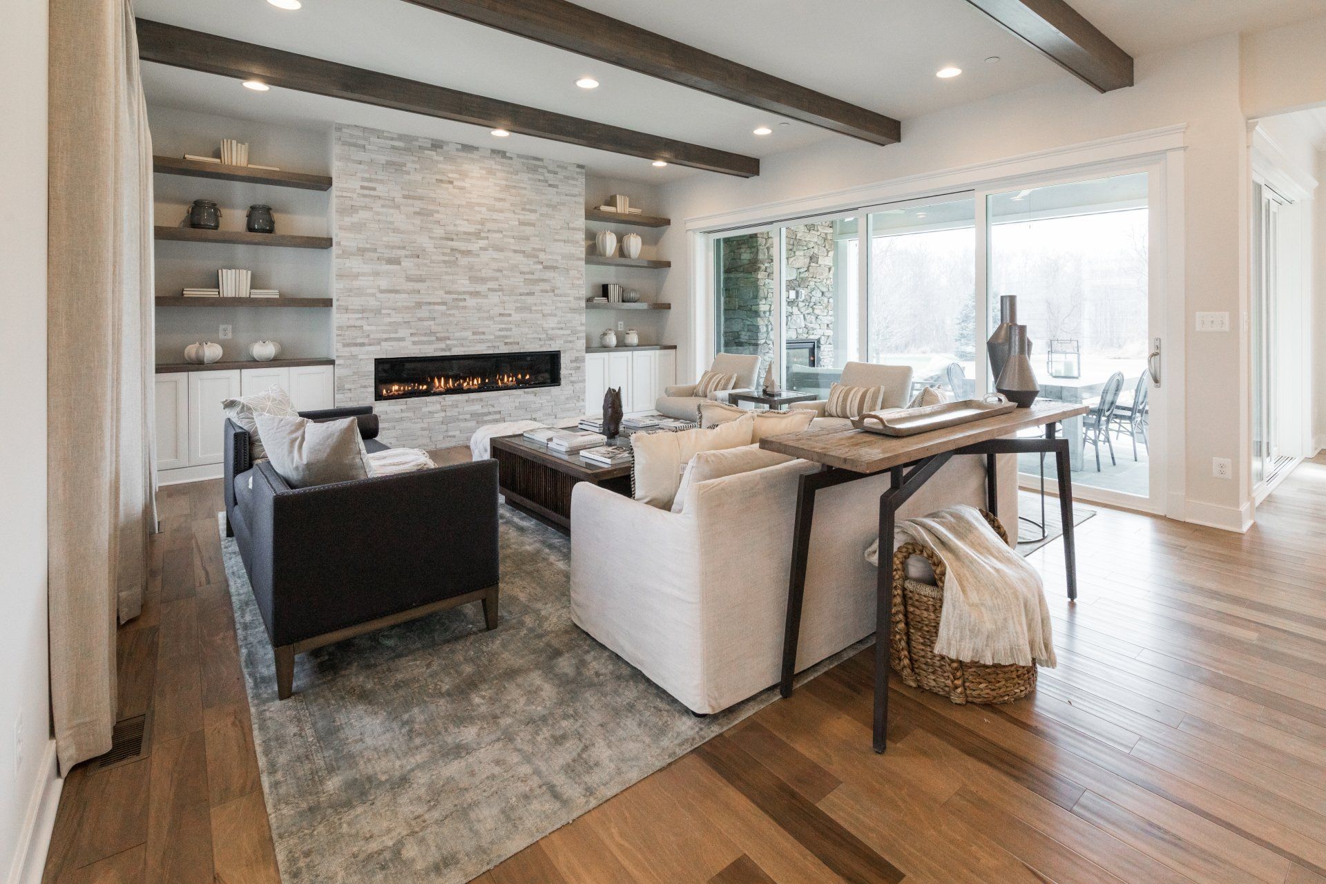 Living room with fireplace, white walls, wood beams, seating, and large windows overlooking a patio.