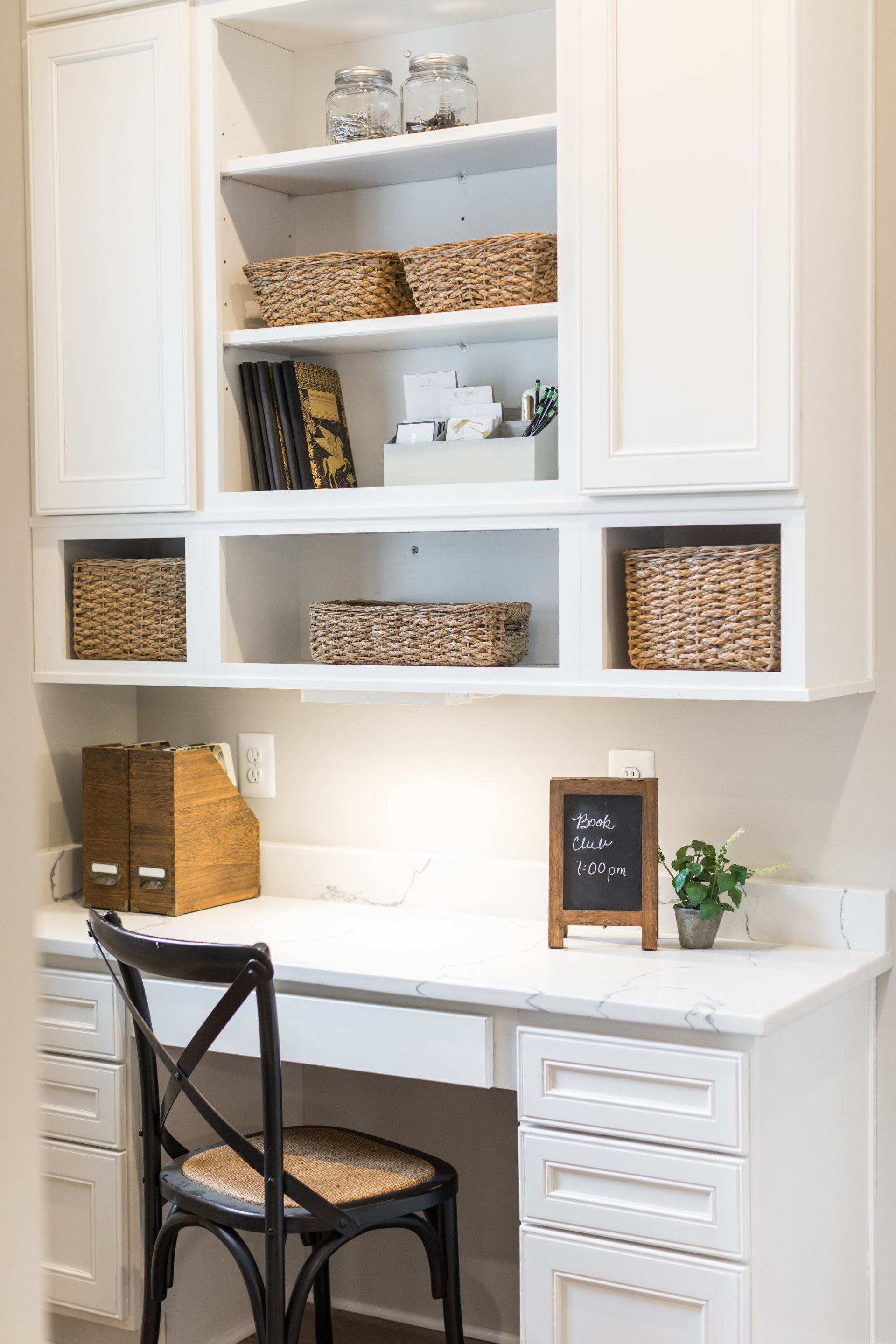 White built-in desk with overhead storage, baskets, and a black chair. Includes a small chalkboard and plant.