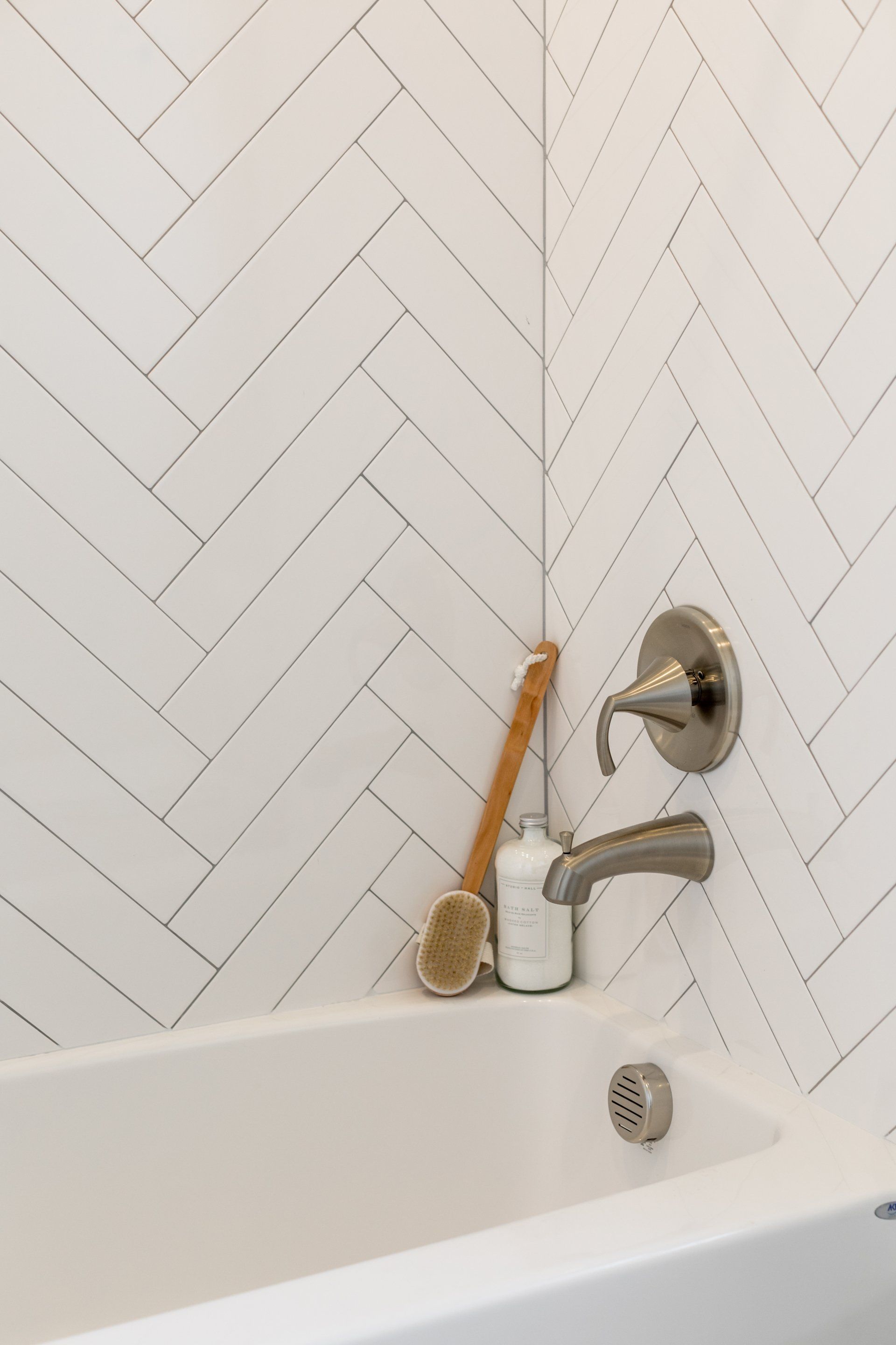 White bathtub with herringbone tile wall, toiletries, and silver faucet.