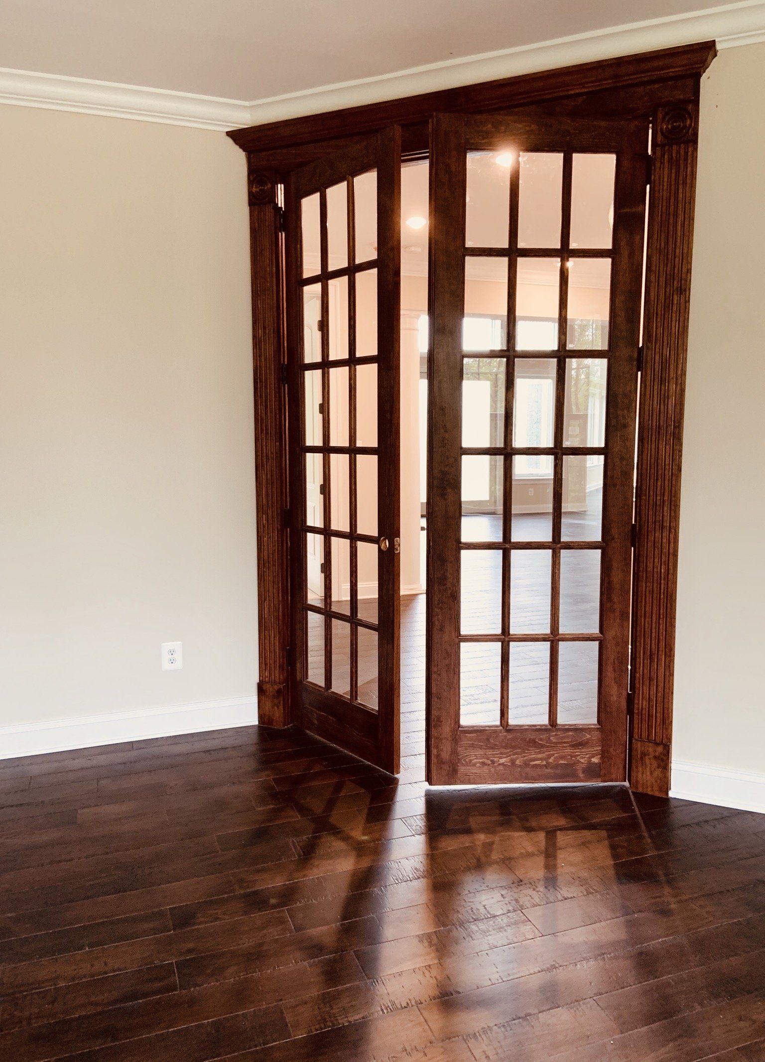 Wooden French doors with ornate trim open to a light-filled room, set in a room with dark wood floors.