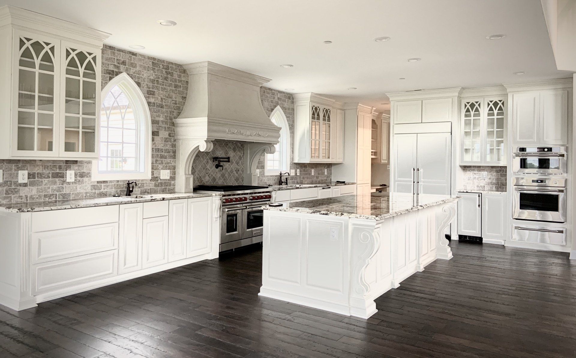 White kitchen with gothic-style cabinets, brick backsplash, stainless appliances, dark wood floor.