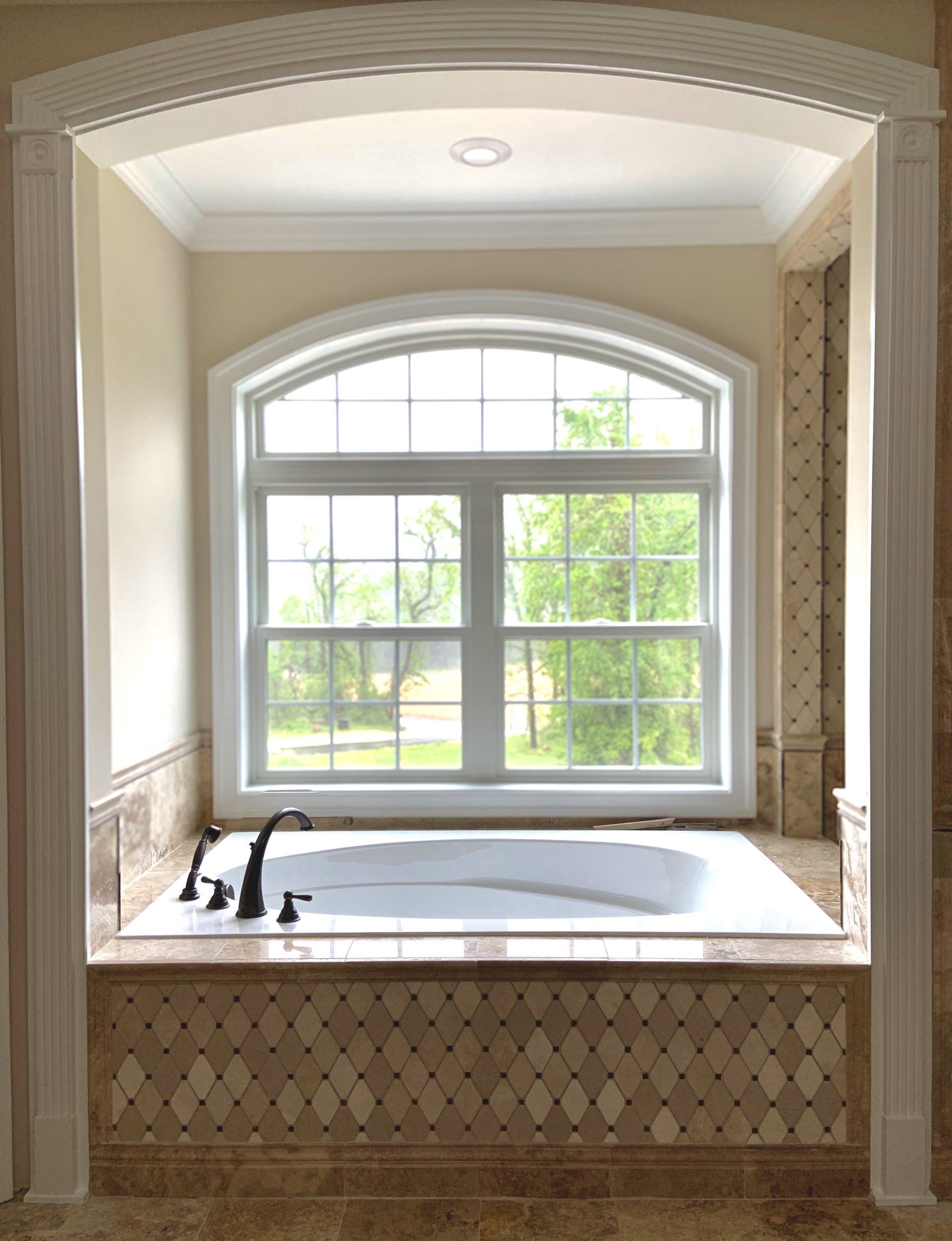 Bathroom with a white bathtub under an arched window, surrounded by tile and white trim.
