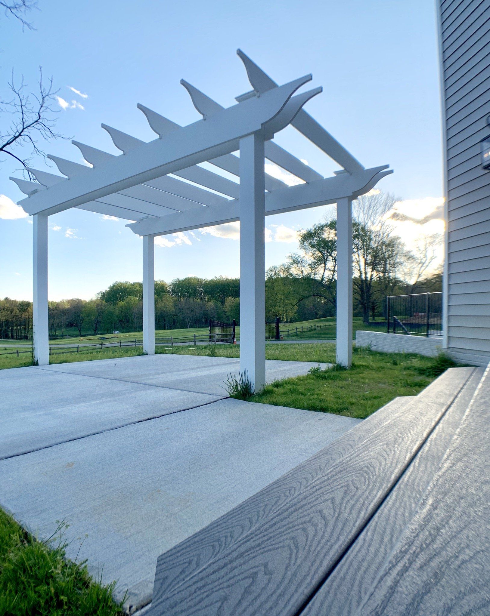 White pergola over concrete patio, next to a house, with green grass and trees under a blue sky.