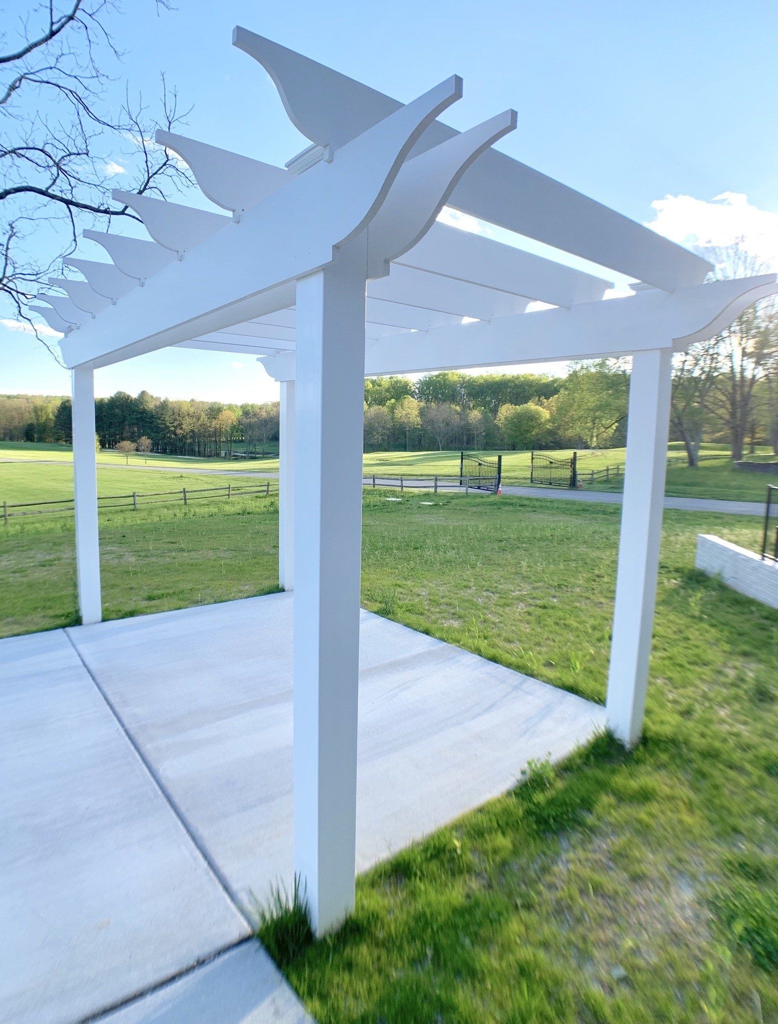White pergola over a concrete patio, in a grassy field on a sunny day.