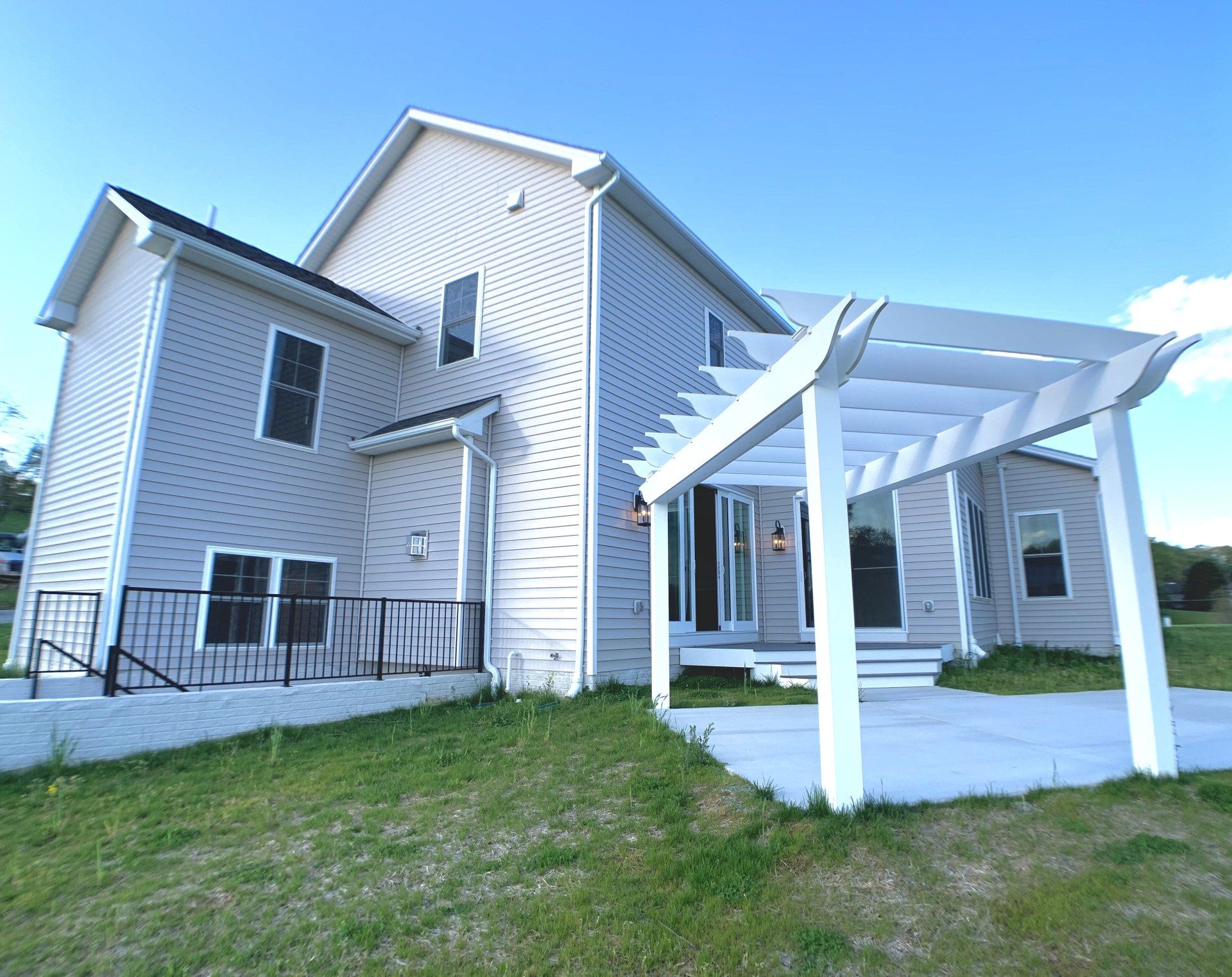 Two-story gray house with white pergola, black railings, and concrete patio on a grassy hill under a blue sky.