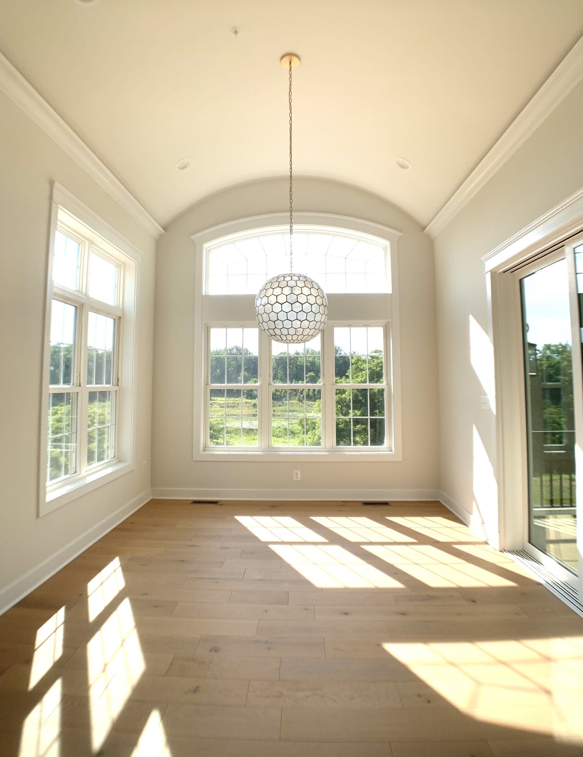 Empty sunlit room with hardwood floors, large windows, and a decorative hanging light.