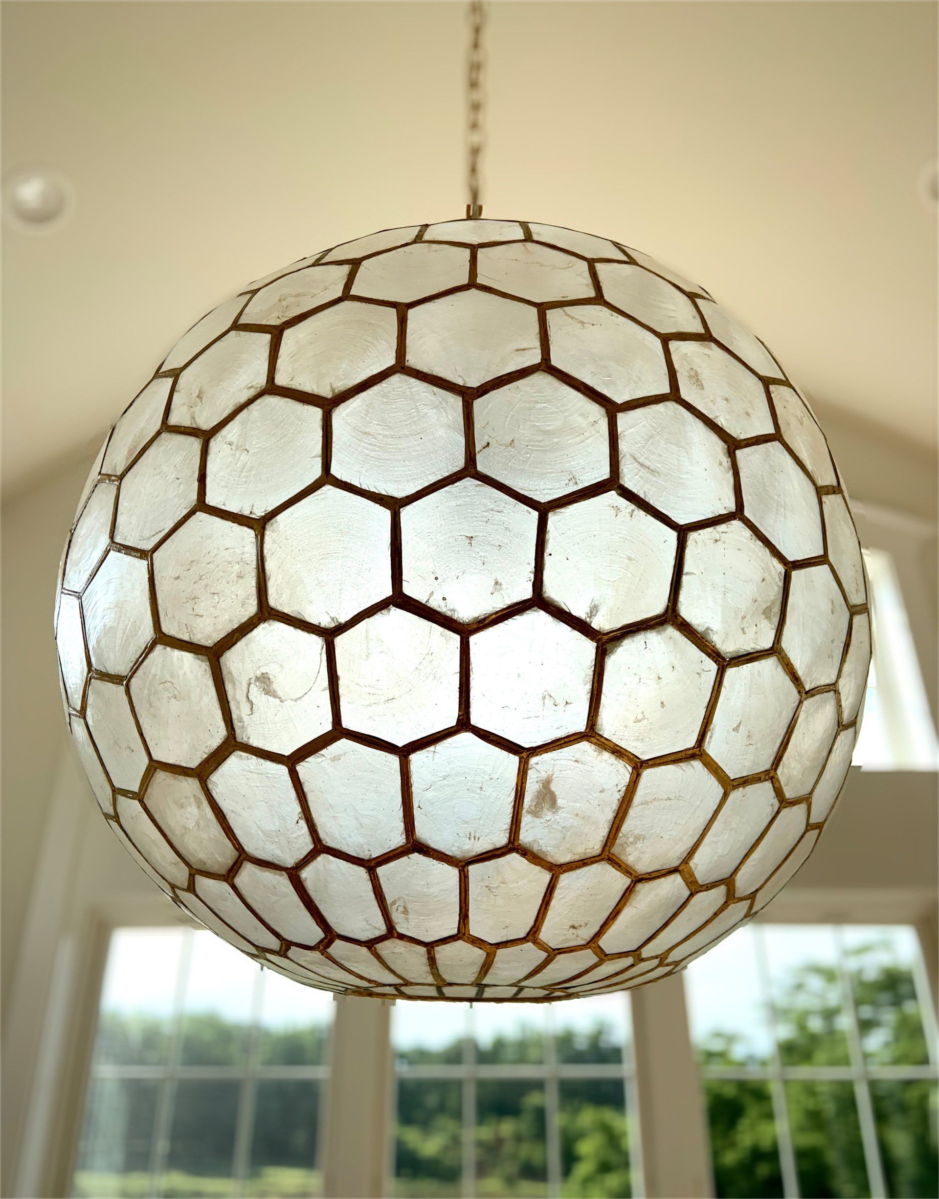Spherical light fixture with honeycomb pattern hanging in a room with windows overlooking a green landscape.
