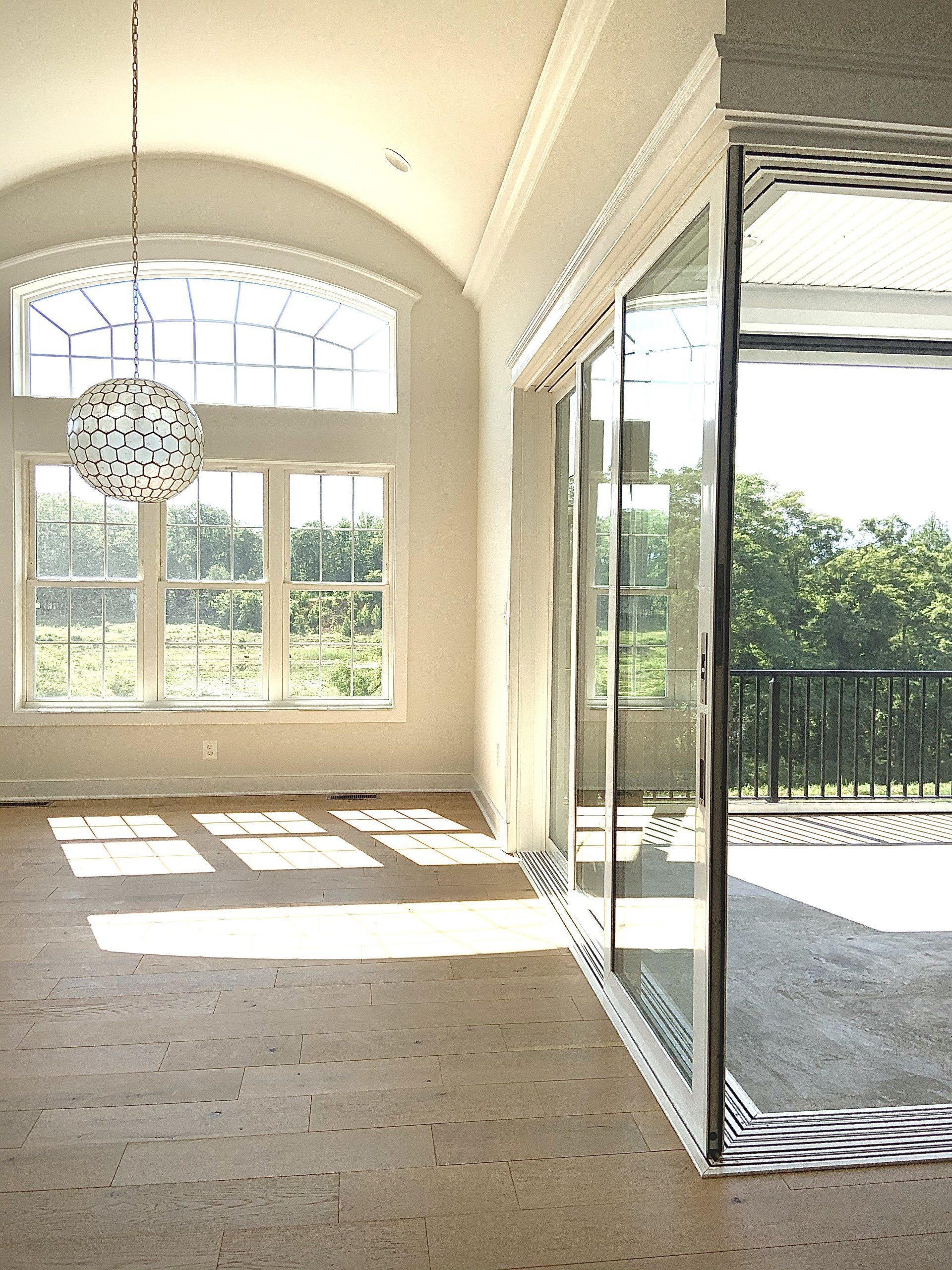 Bright, empty room with large windows, sliding glass doors, and decorative light fixture; view of trees outside.