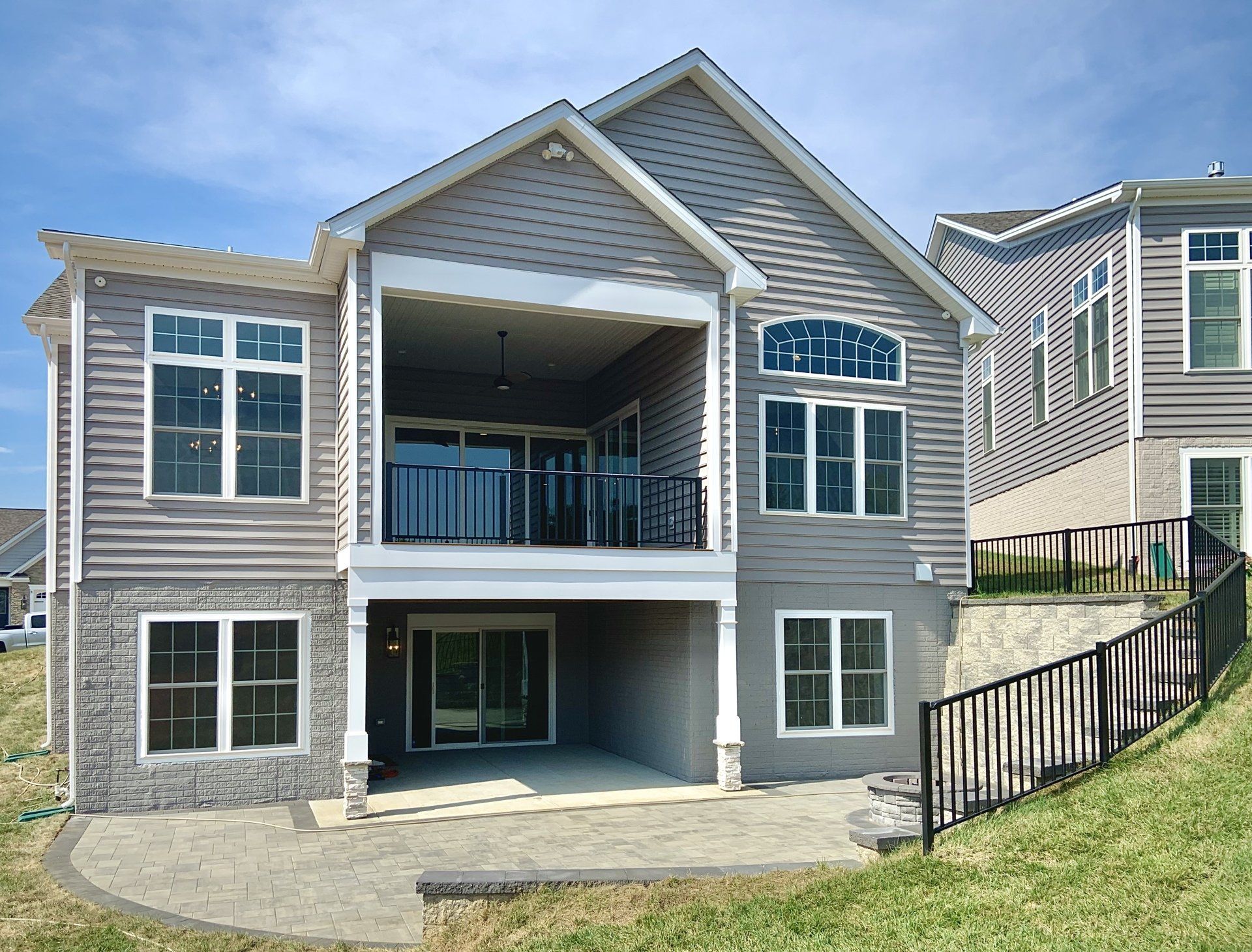 Rear view of a gray house with a balcony and patio on a grassy hillside under a blue sky.