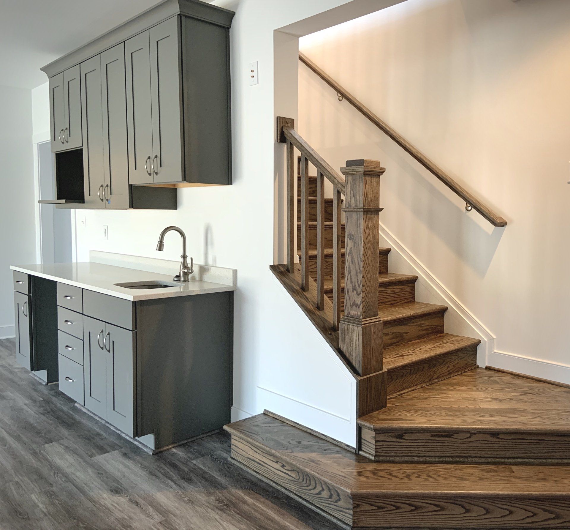 Gray cabinets and wet bar next to a wooden staircase with handrail in a bright home.