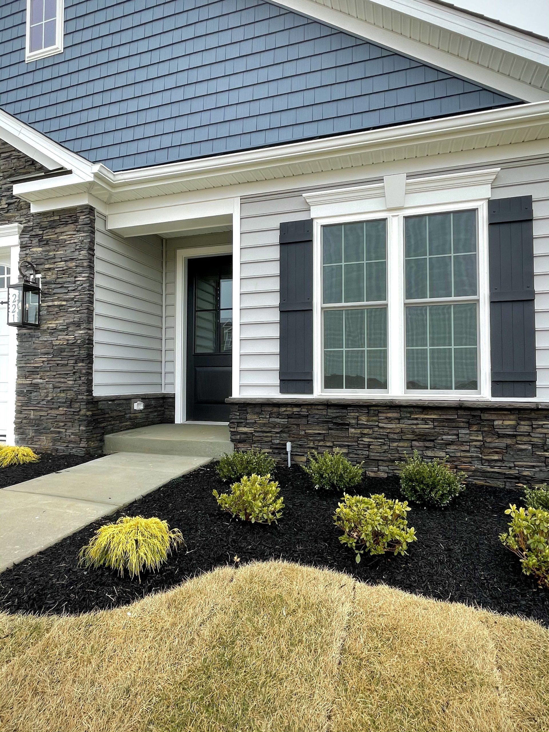 Exterior view of a house with blue siding, stone accents, dark shutters, and a walkway, with landscaping.