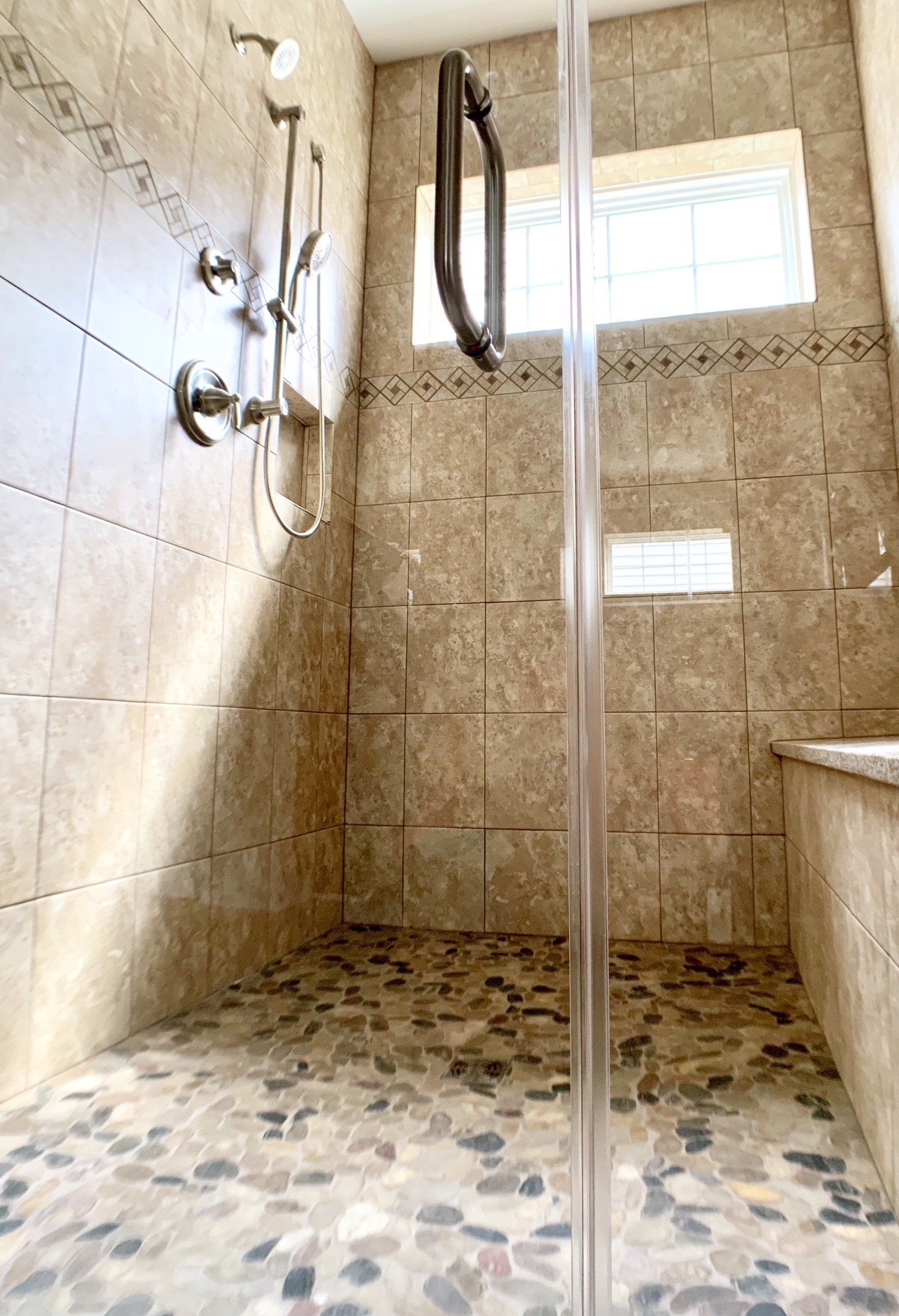 Shower with stone floor and beige tile walls, glass door, and silver fixtures.