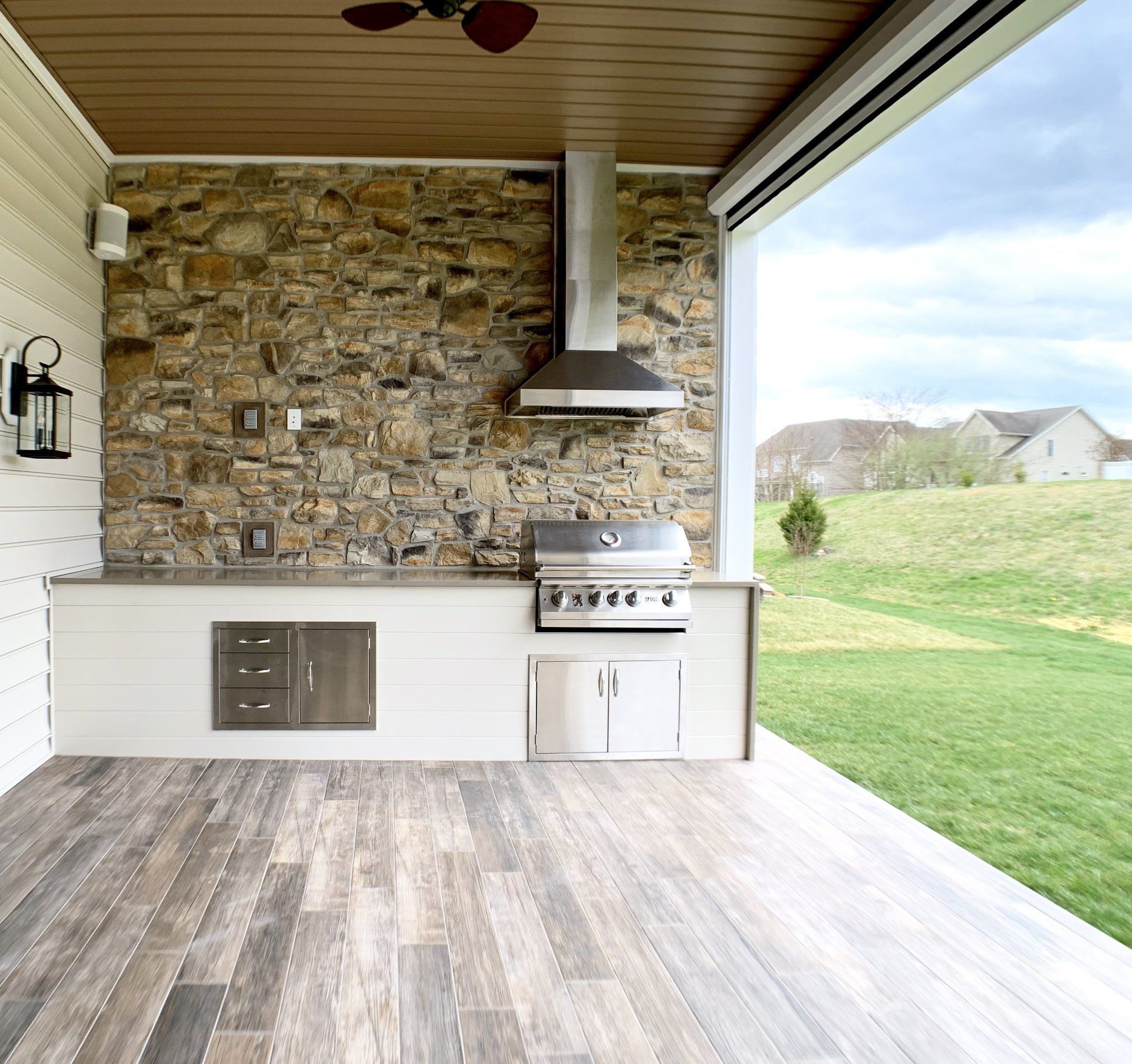 Outdoor kitchen with stone wall, grill, and wooden floors.