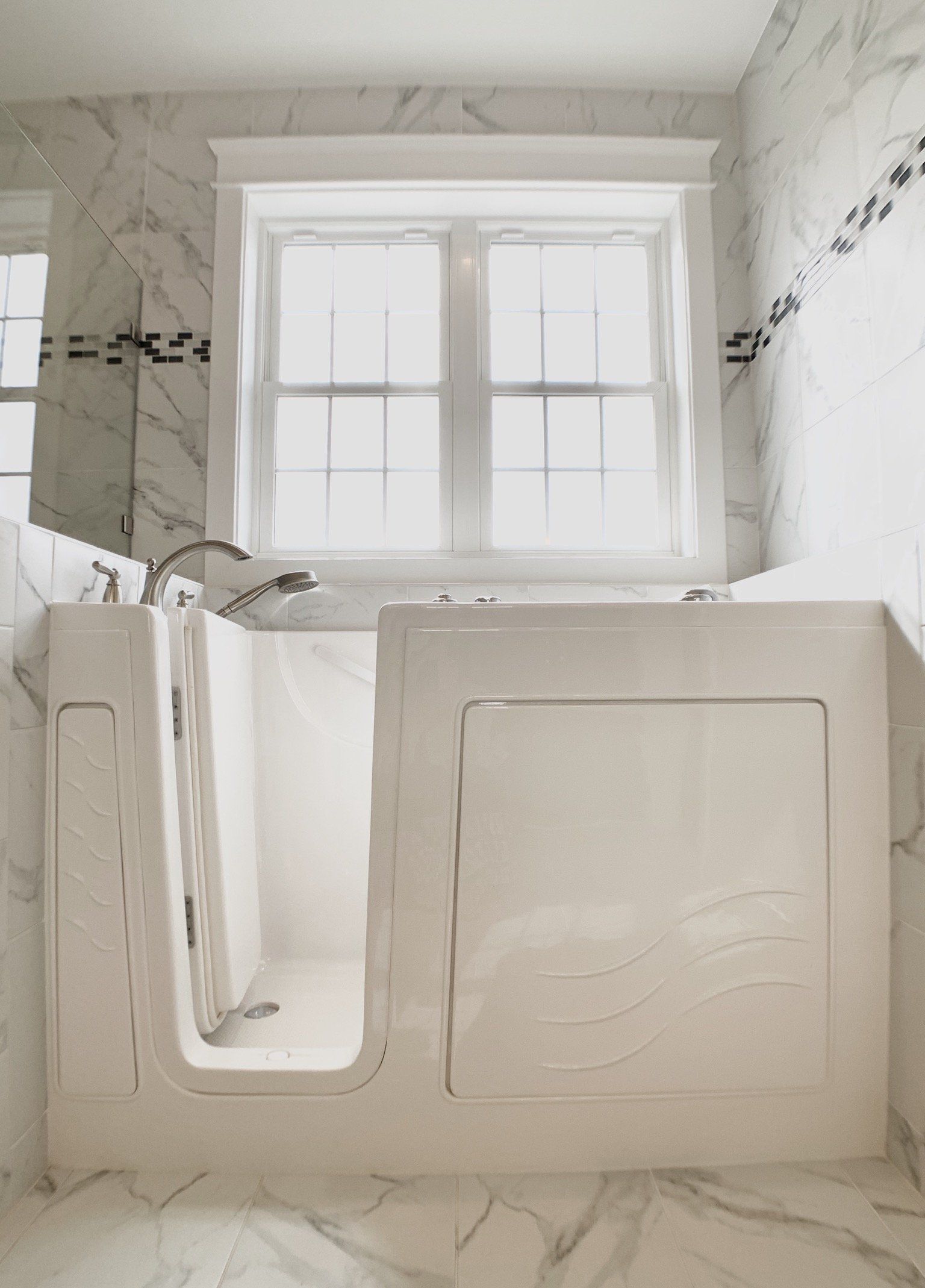 White walk-in bathtub under a window in a marble-tiled bathroom.