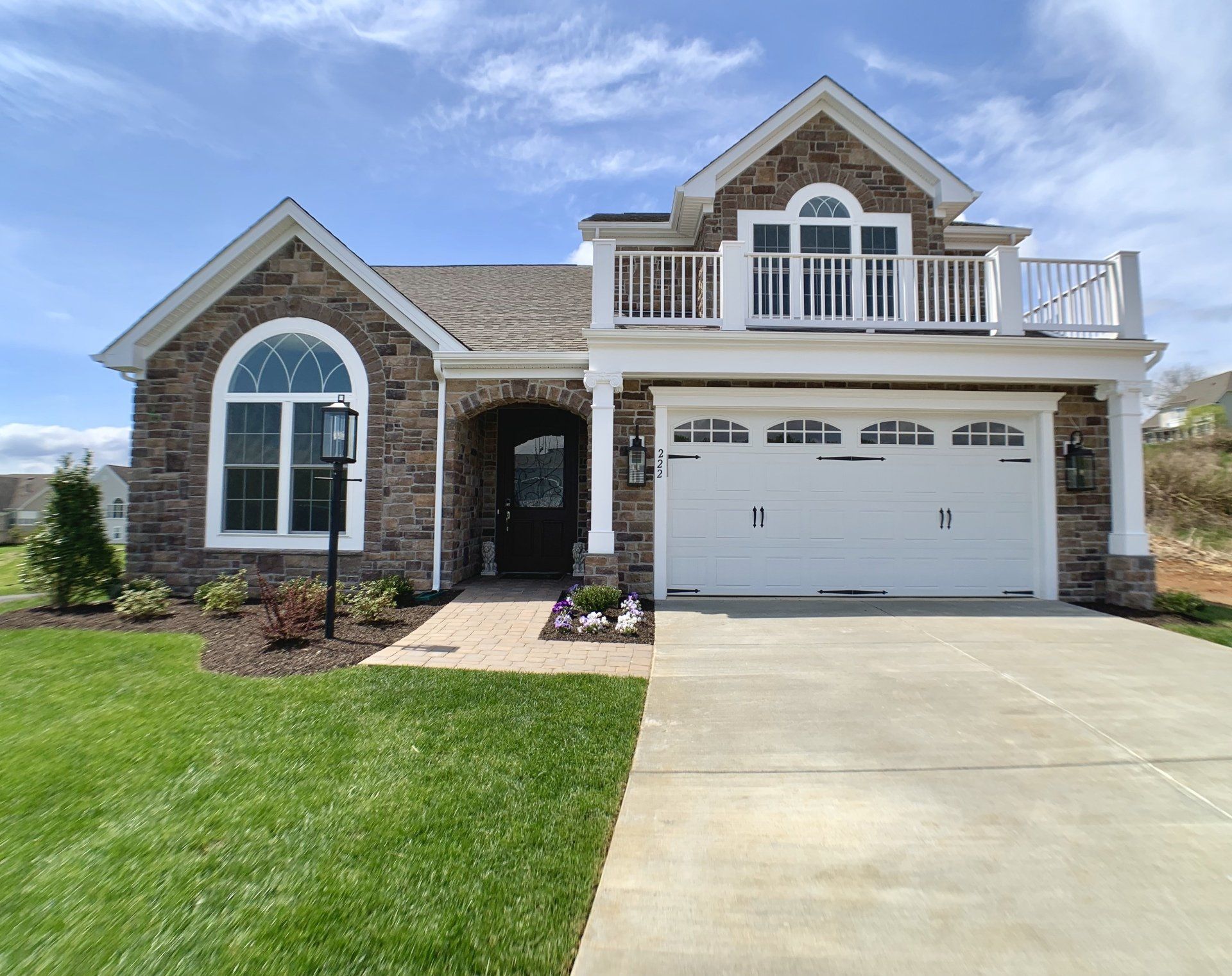 House with stone exterior, arched windows, and a balcony. Garage door, green lawn, and blue sky.