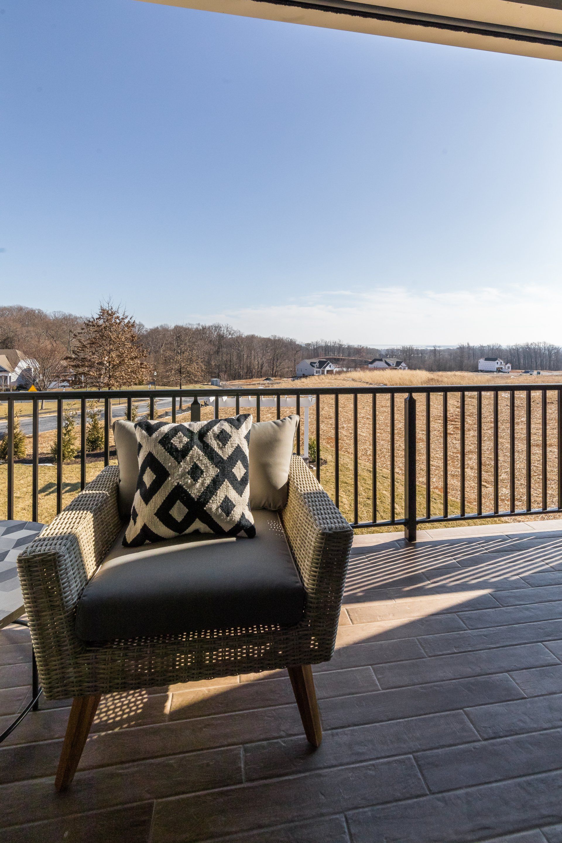 Wicker chair with pillows on a patio overlooking a field, under a blue sky.