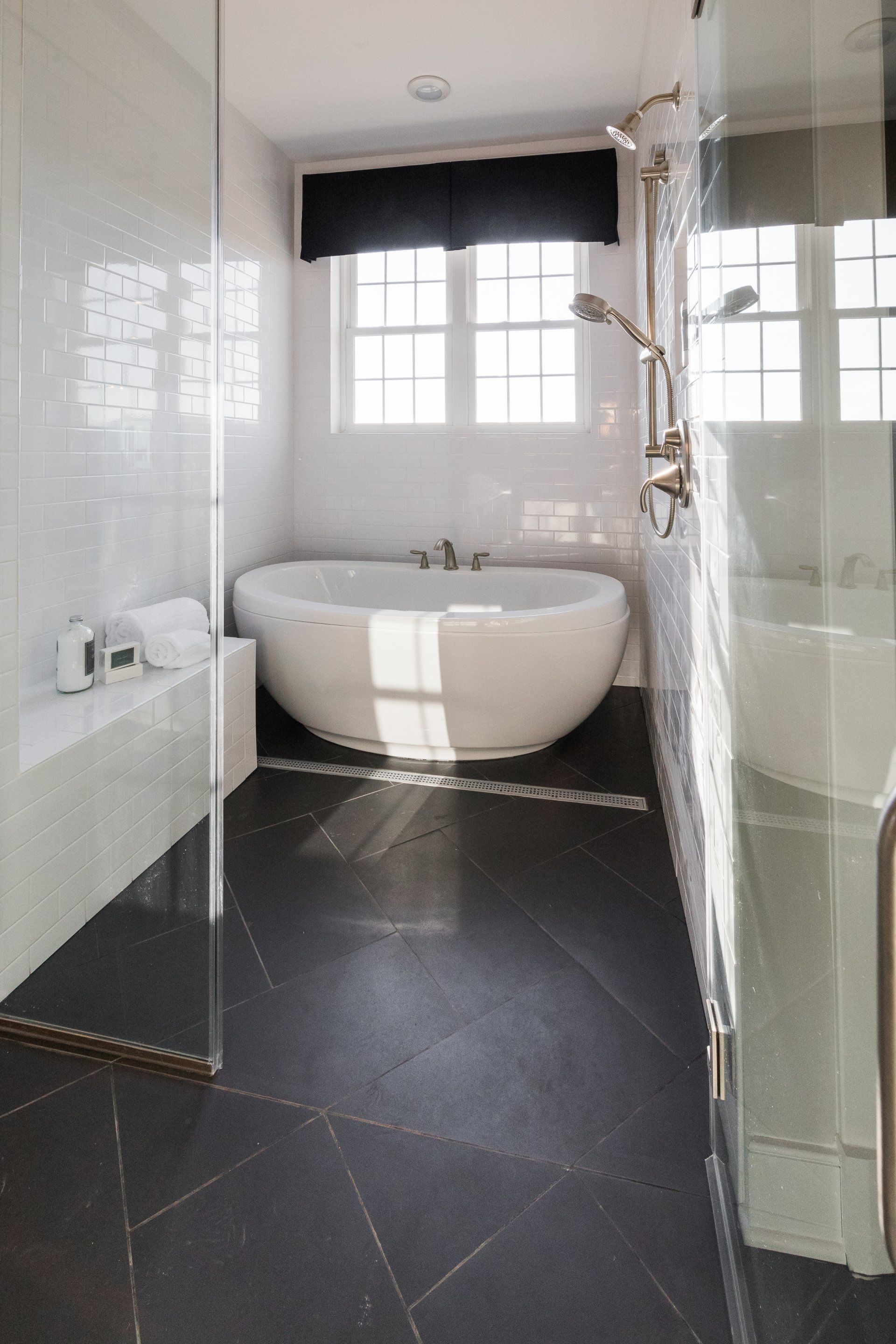 Modern bathroom with a white tub under a window, dark tile floor, and a glass shower.