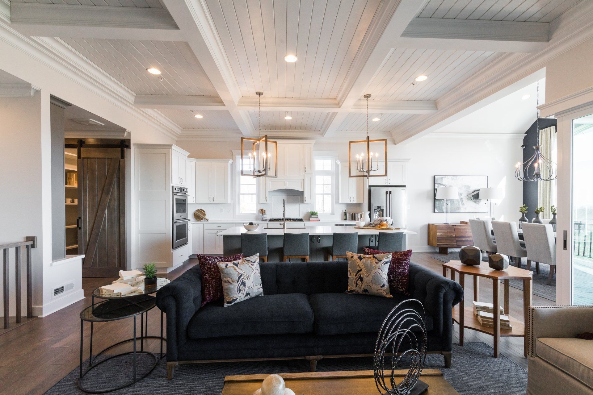 Living room with a dark sofa, white kitchen, and wood ceiling beams.