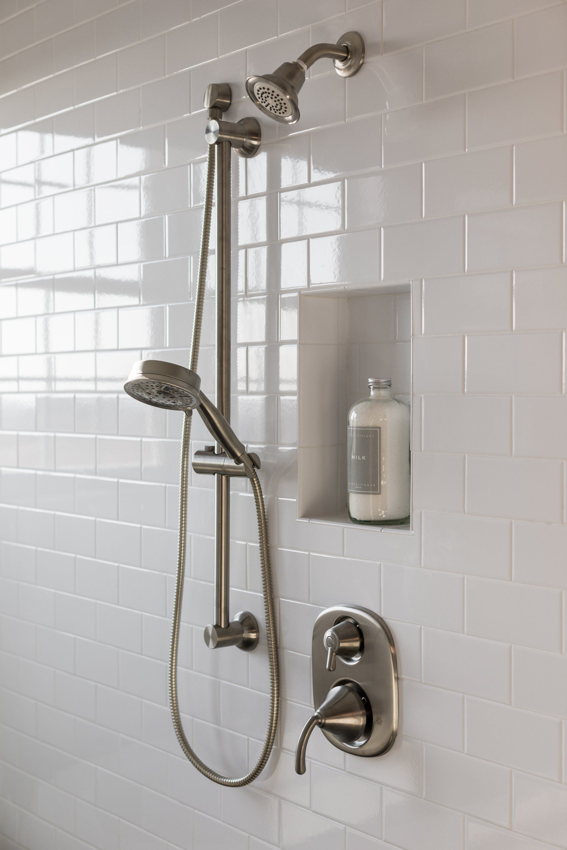 White-tiled shower with silver fixtures, including a handheld showerhead, fixed showerhead, and soap niche.
