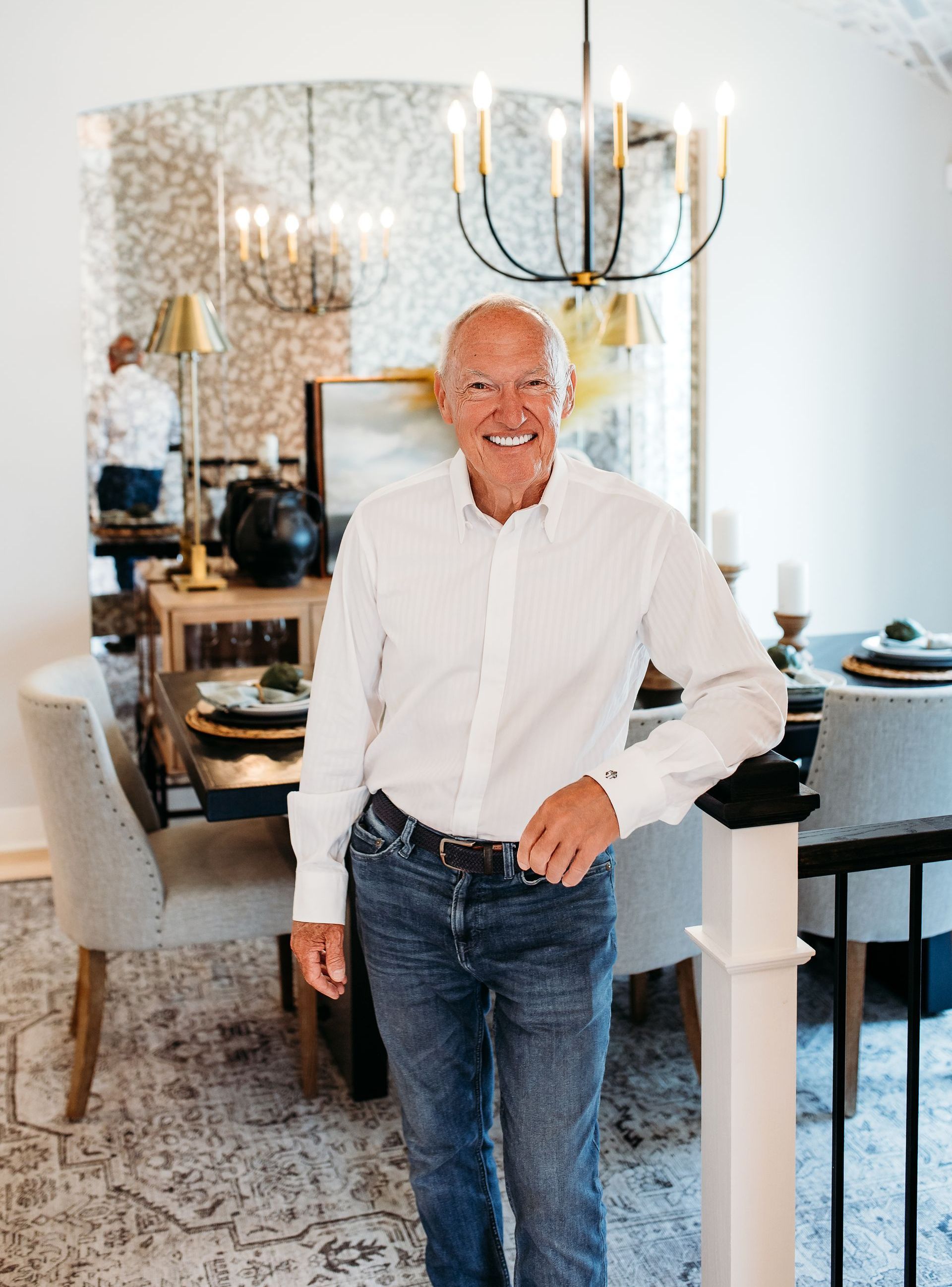 Man in white shirt and jeans smiles, leaning on a railing, in a dining room setting.