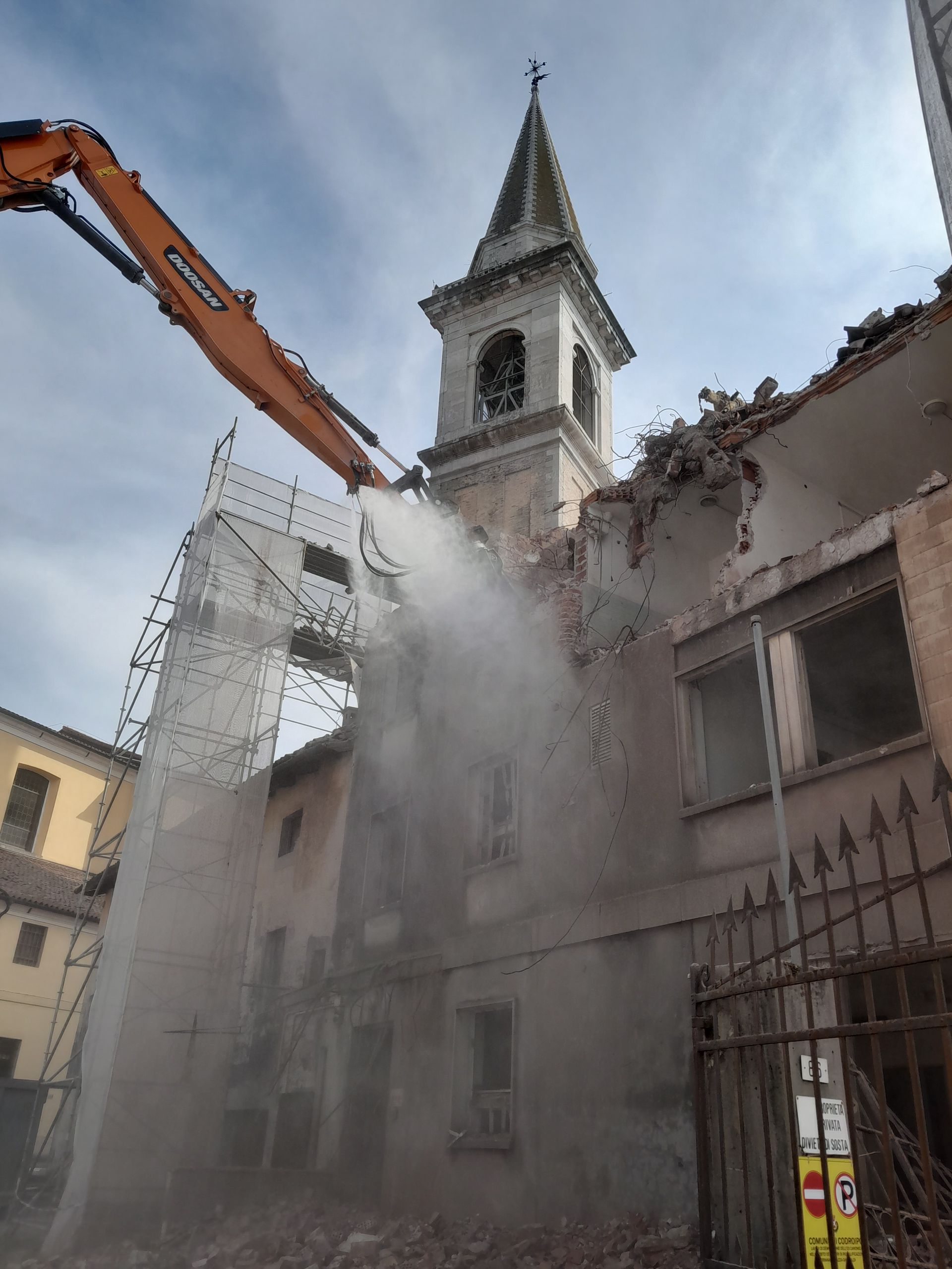 Una gru sta demolendo un edificio con una torre dell'orologio sullo sfondo.