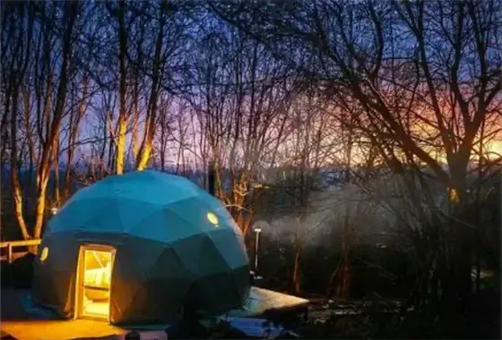 Blauwe geodetische koepel, gloeiende deuropening, in een bosrijk gebied bij schemering; rook stijgt op uit de schoorsteen.