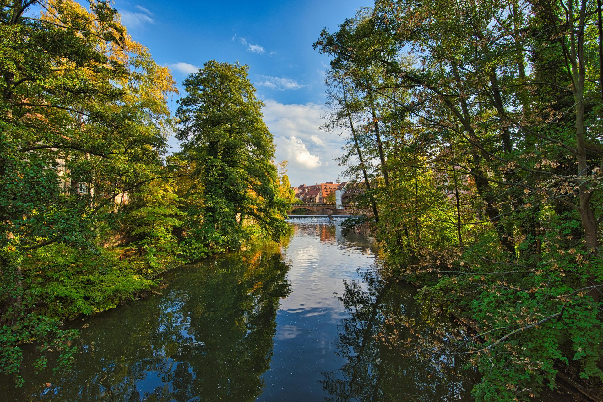 Een kalme rivier stroomt door een weelderig groen bos en weerspiegelt de lucht en de gebouwen in de verte.