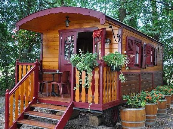 Houten caravan met bordeauxrode accenten, een kleine veranda, potplanten en een trap.