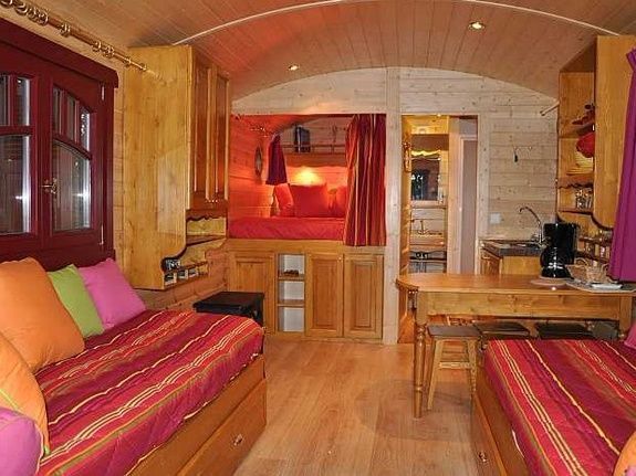 Gezellig interieur van een woonwagen met houten wanden, rood en oranje beddengoed en een kleine eettafel.