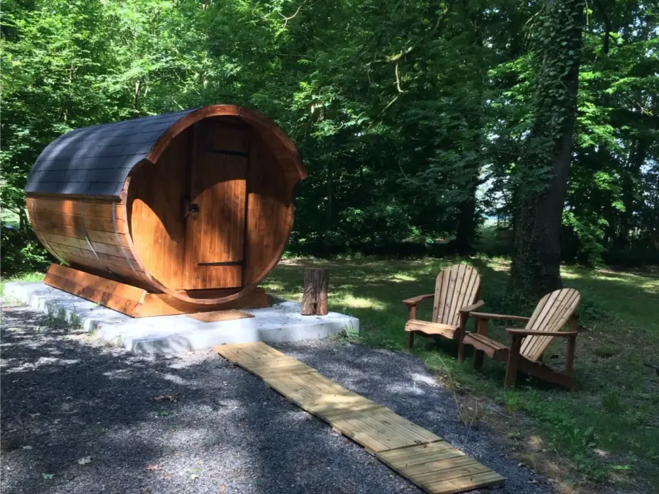 Tonvormige sauna met een houten deur, omgeven door bomen, met twee stoelen. Een houten pad leidt naar de sauna.