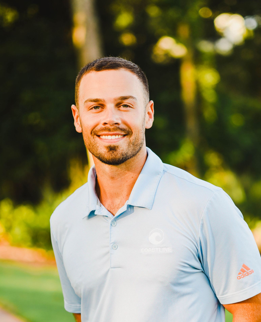 A smiling person in a light blue polo shirt stands outdoors with a softly blurred, warm-toned, leafy background.