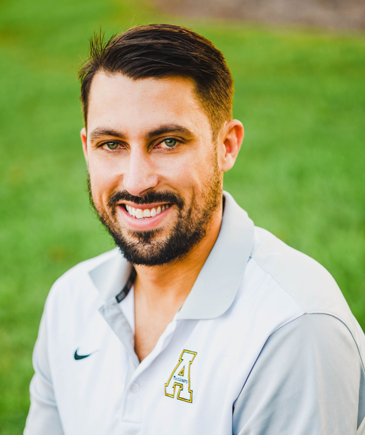 A smiling person with a beard, wearing a white polo shirt with an emblem, outdoors against a blurred green background.