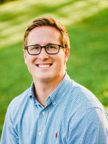 A person wearing glasses and a blue gingham shirt smiling outdoors with a green, blurred background.