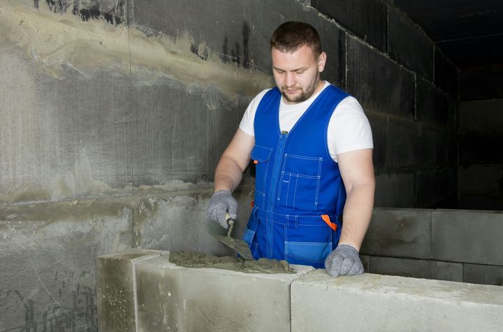 Construction worker in blue overalls laying concrete blocks in a dim unfinished room