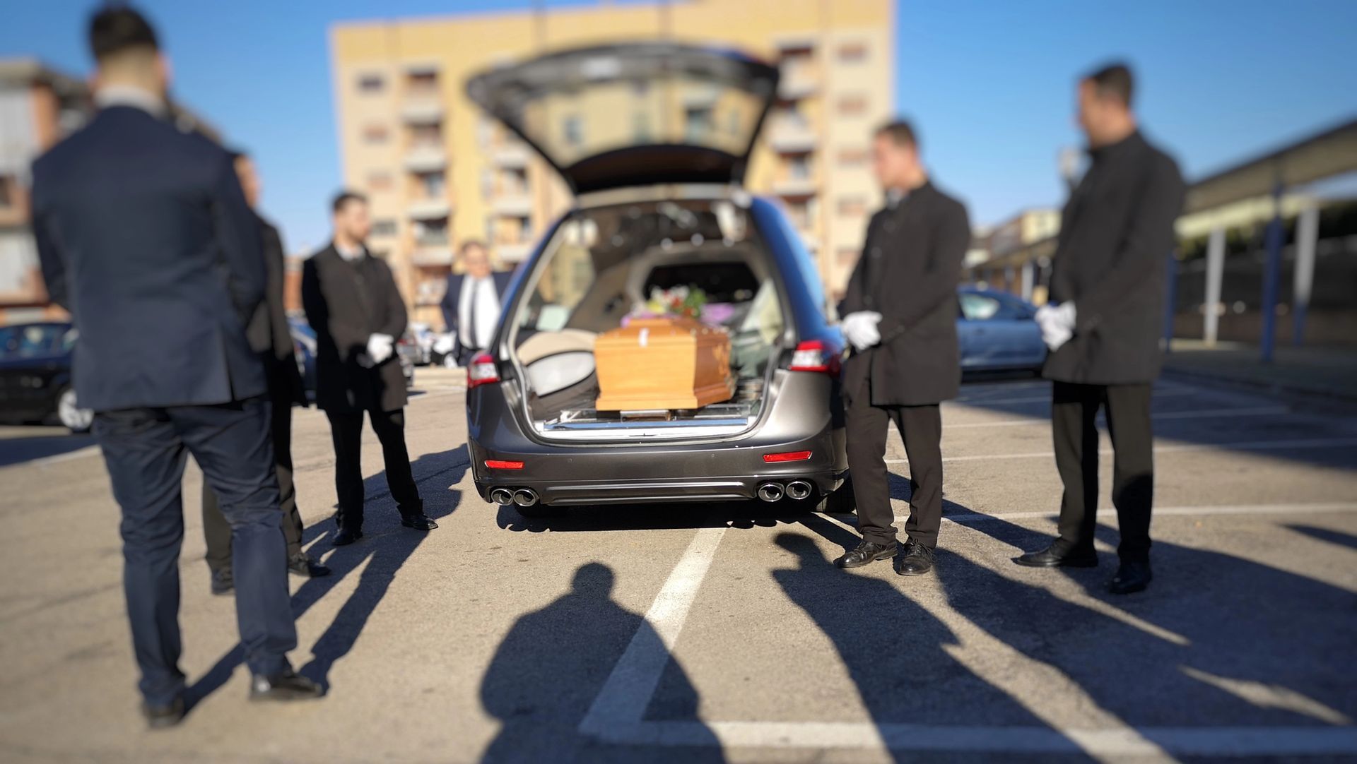 Carro funebre con bagagliaio aperto; i portatori della bara circondano una bara in un parcheggio in una giornata di sole.