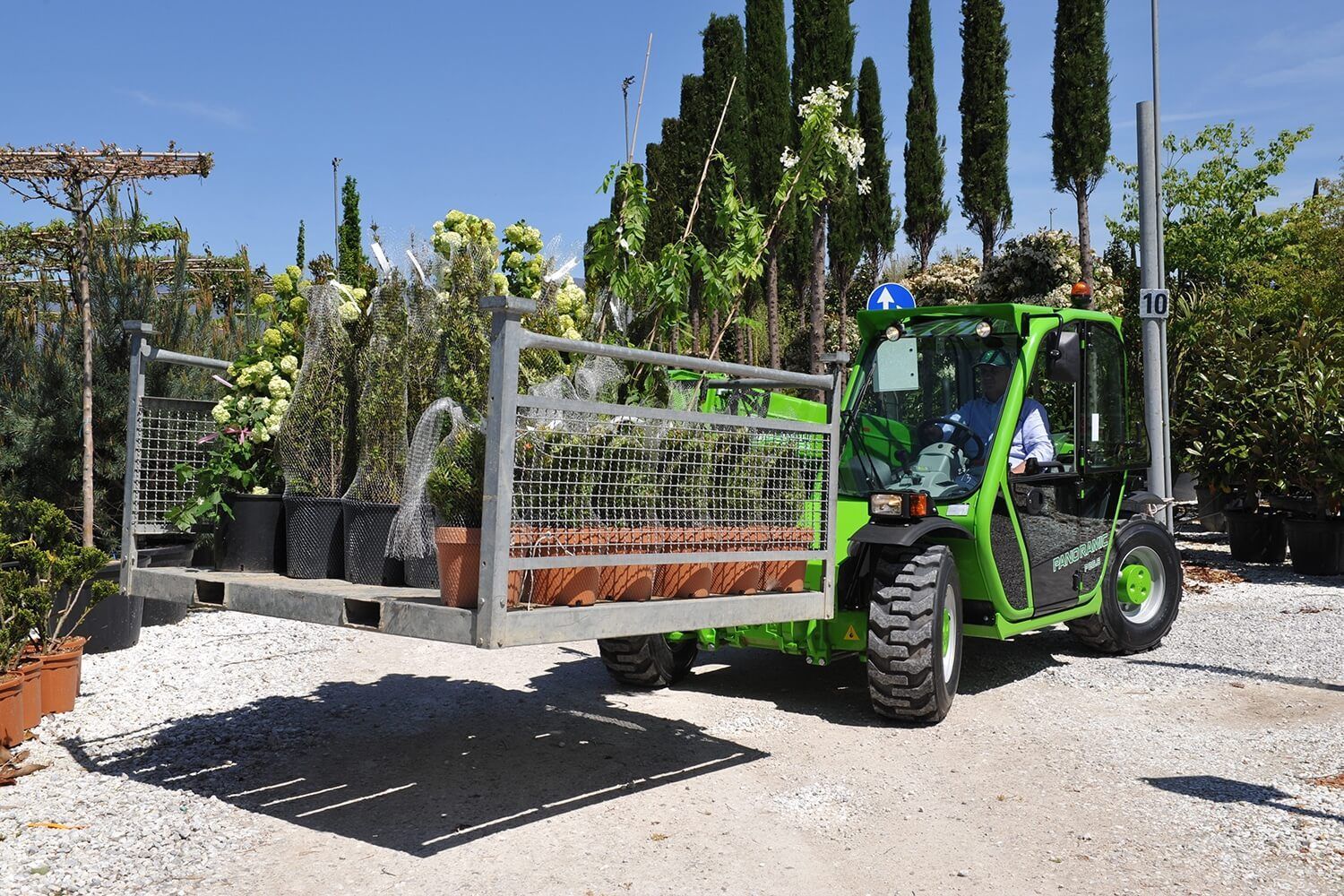Un chariot élévateur vert transporte une caisse de plantes en pot dans un jardin.