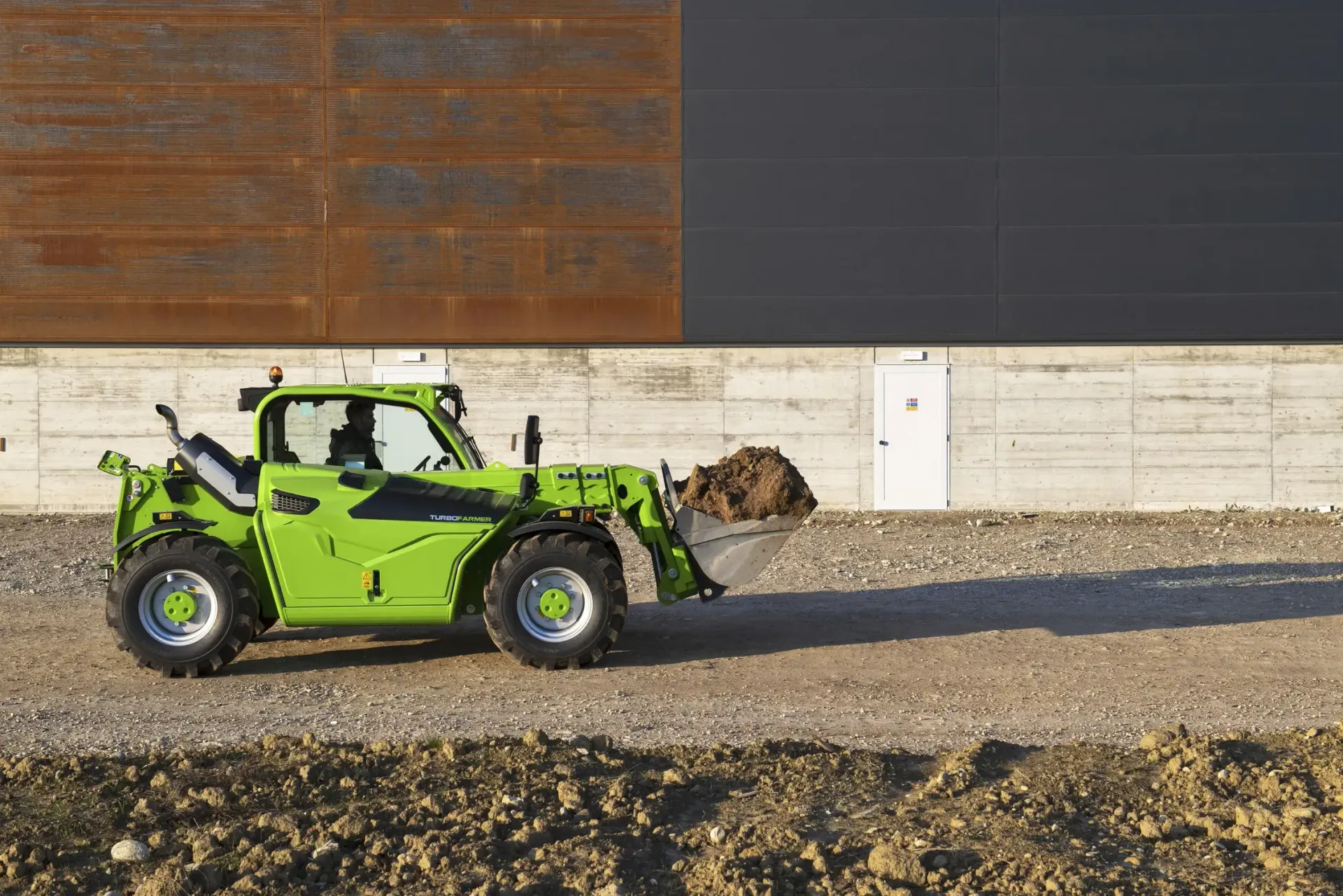 Un chariot élévateur vert roule sur un chemin de terre devant un bâtiment.