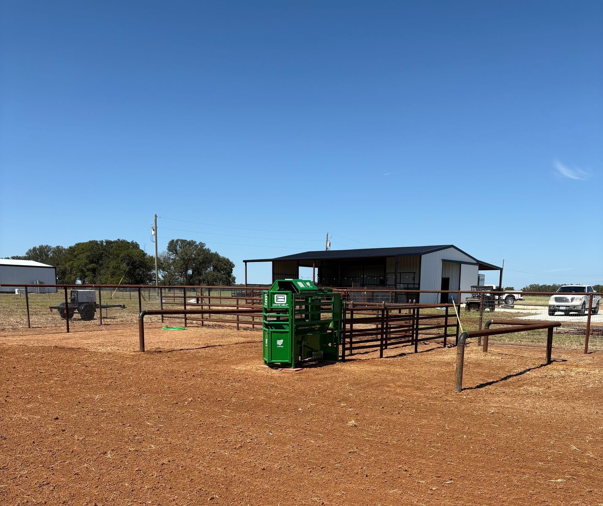 Cattle working area with green chute, brown fences, and a metal building under a blue sky.