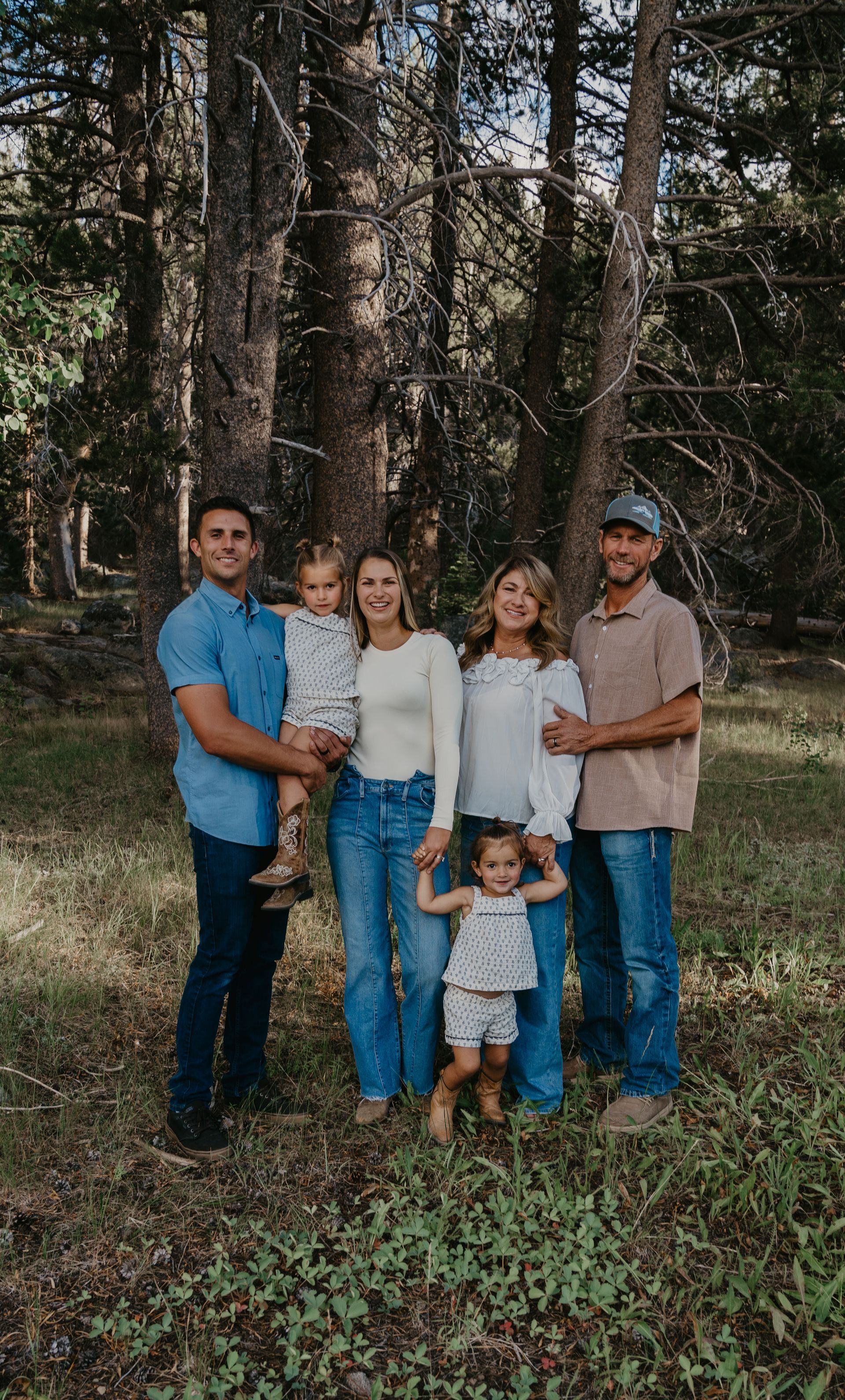 Family of six standing in a forest. Adults wearing jeans and button-down shirts, children in summer outfits.