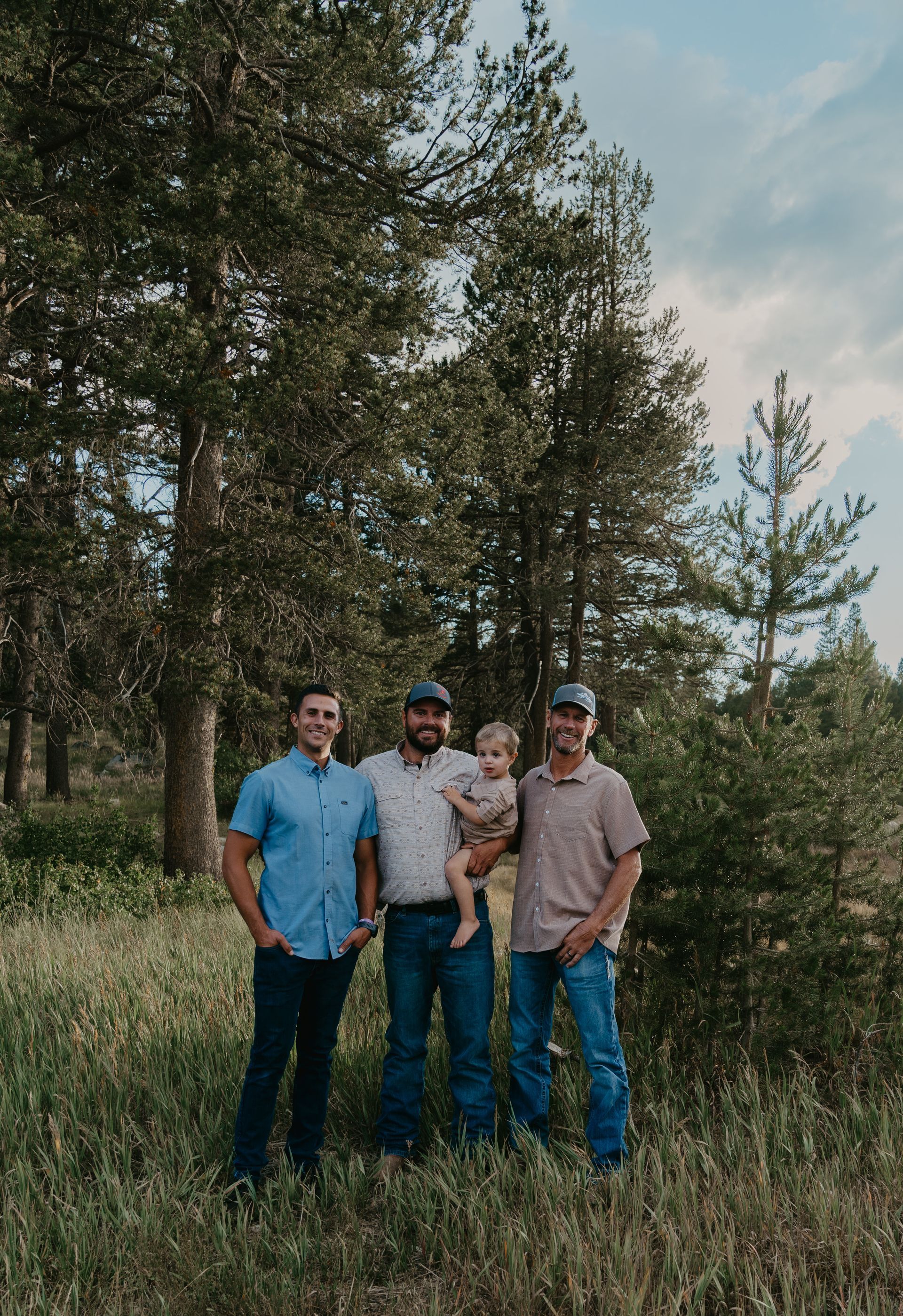 Four people standing in a wooded area. One holds a young child. All are wearing casual clothing.