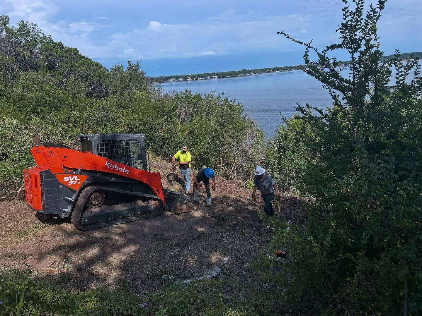 Orange skid steer and workers clearing brush near water with a bridge in the distance.