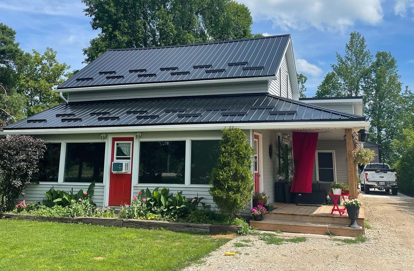 A light-colored house with a dark metal roof, red door, and front porch, surrounded by green landscaping and trees.