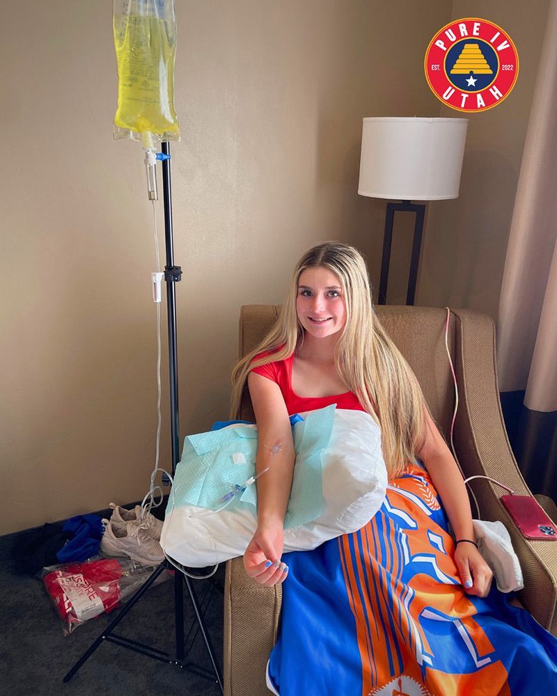 A young woman smiles while receiving an IV hydration drip infusion on a chair in a hotel room for quick recovery or wellness.