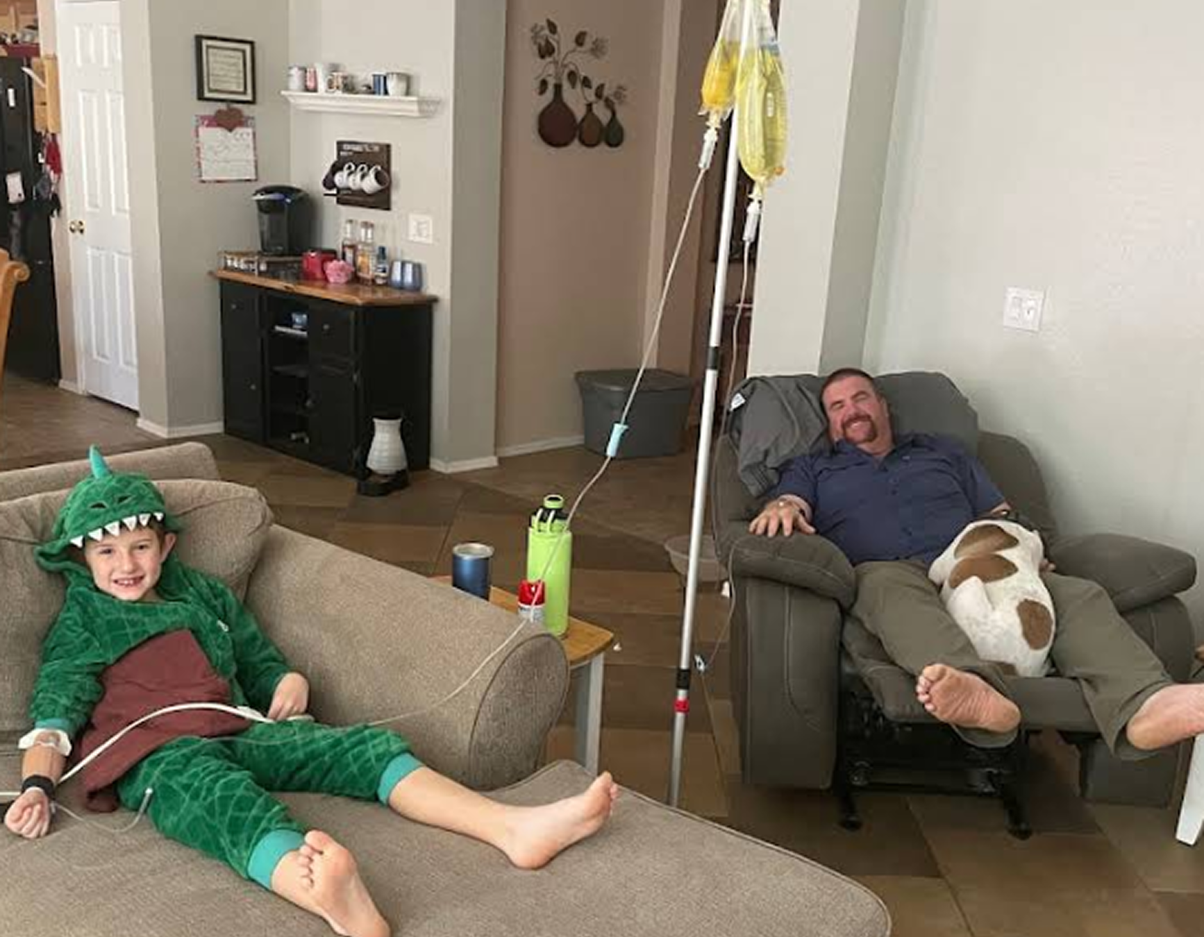 A boy in a dinosaur costume is laying on a couch next to a man in a chair.